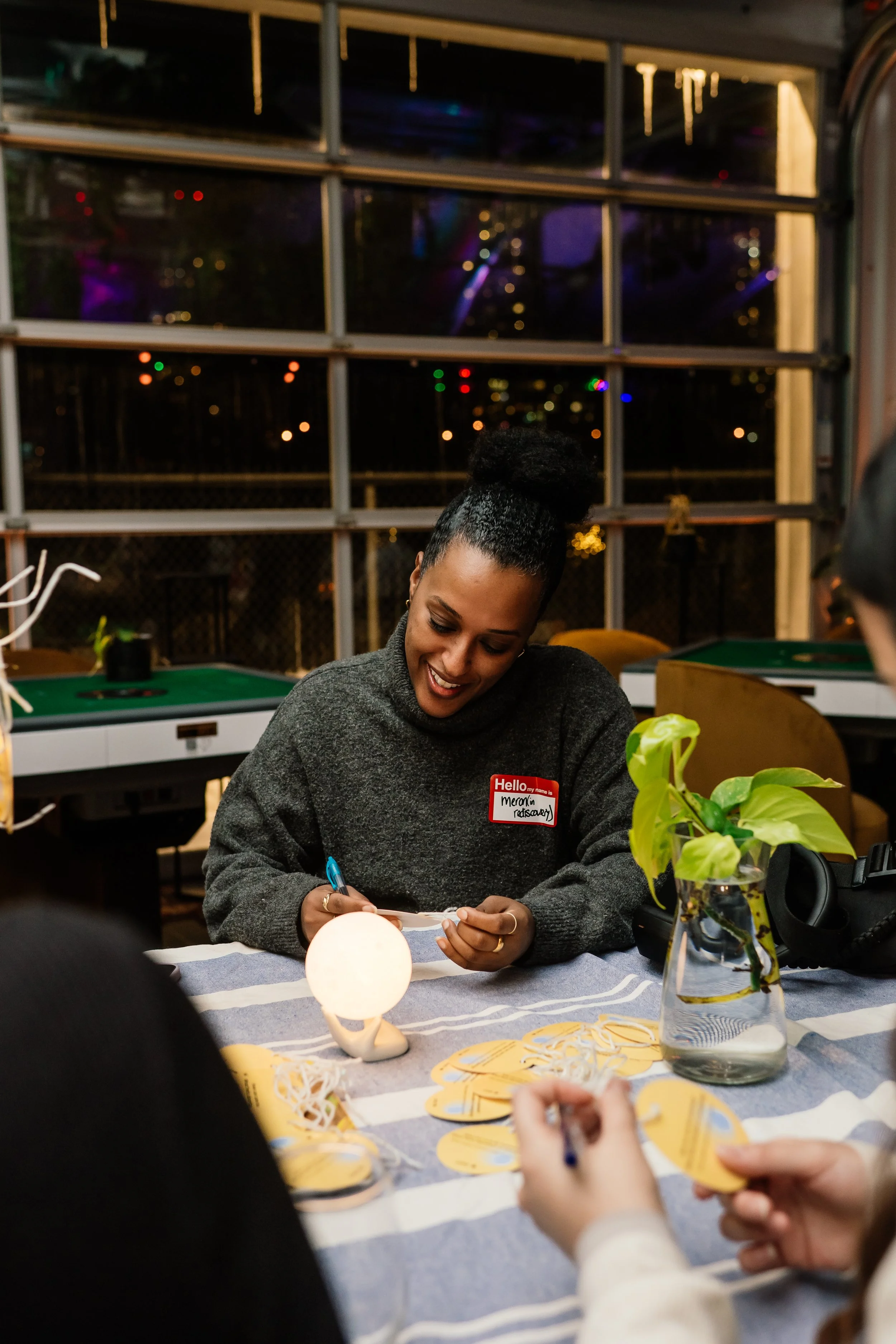 A woman with a name tag that reads 'Hello my name is Meron' is smiling while sitting at a table with a group of people. There are cards or tags on the table, a small glowing lamp, and a potted plant in a vase. The setting appears to be indoors with windows and colorful lights visible in the background.