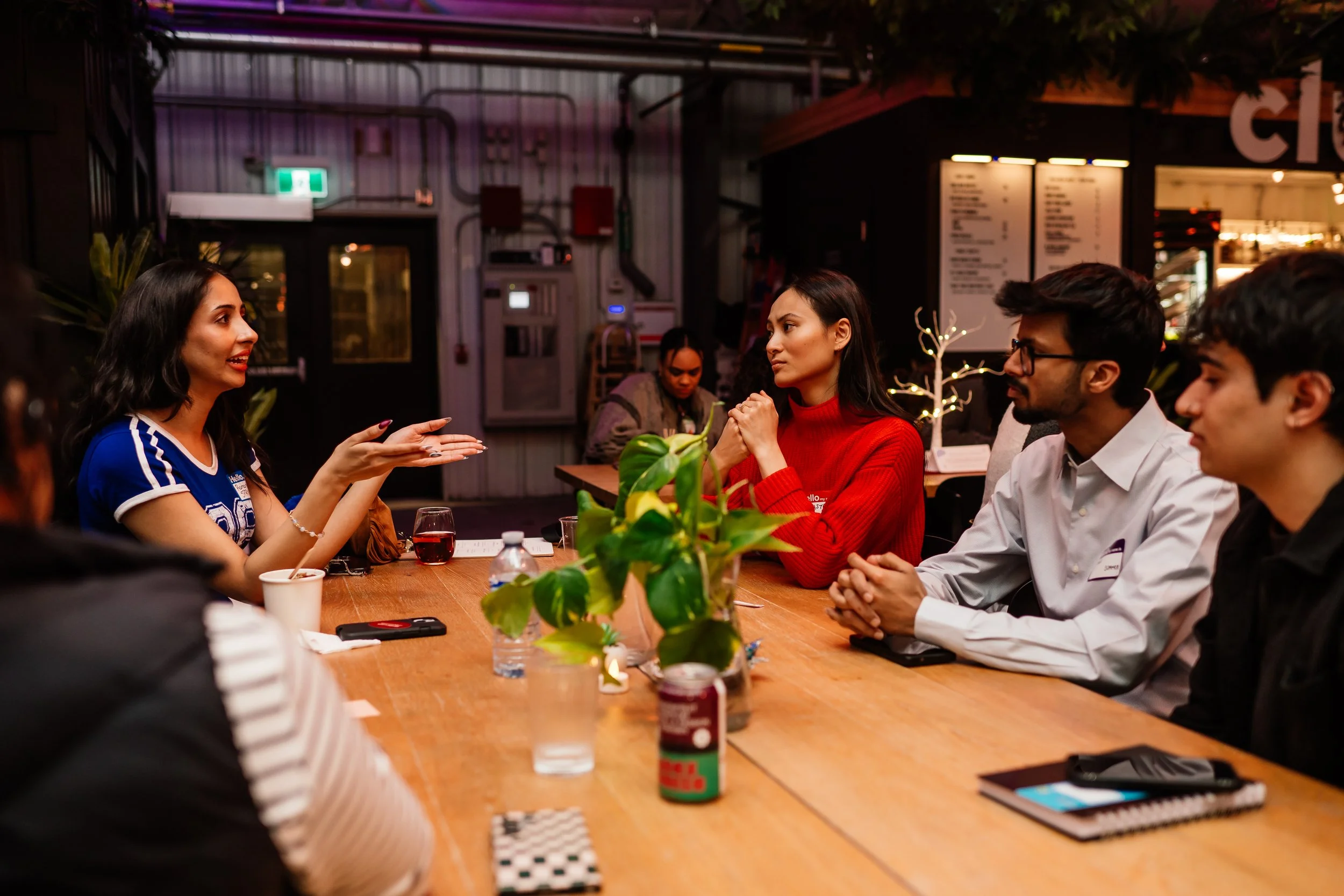 A group of six young adults are sitting around a wooden table in a dimly lit restaurant, engaged in a conversation. The woman on the left is wearing a blue sports jersey and is gesturing with her hands, while the woman in the red sweater listens attentively. Other members are sitting beside her, with some having notebooks and drinks on the table, and a small decorative plant in the center.