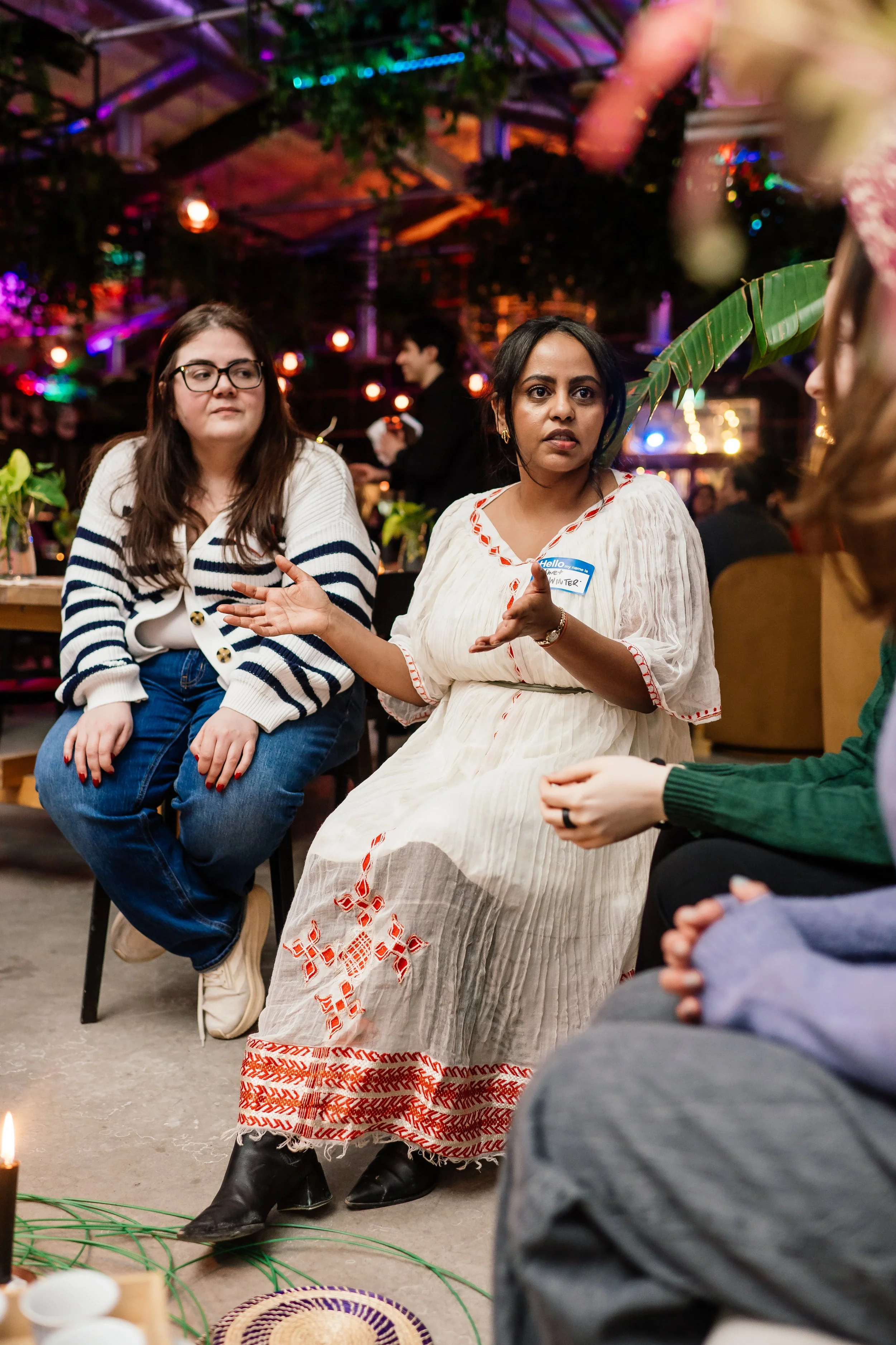A group of women sitting and engaging in a conversation in a colorful, decorated indoor setting with string lights and plants.