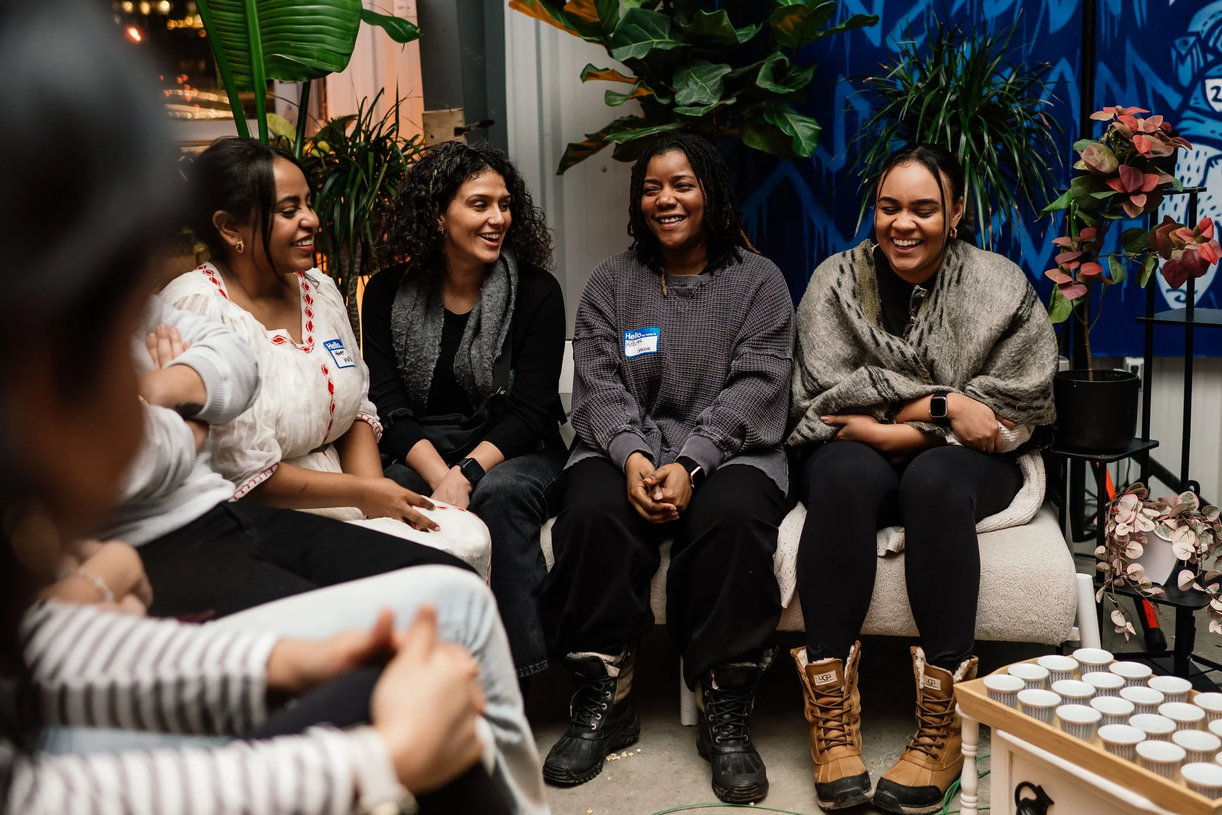 Group of five women sitting closely together, smiling, and enjoying conversation in an indoor setting with plants and colorful wall art in the background.