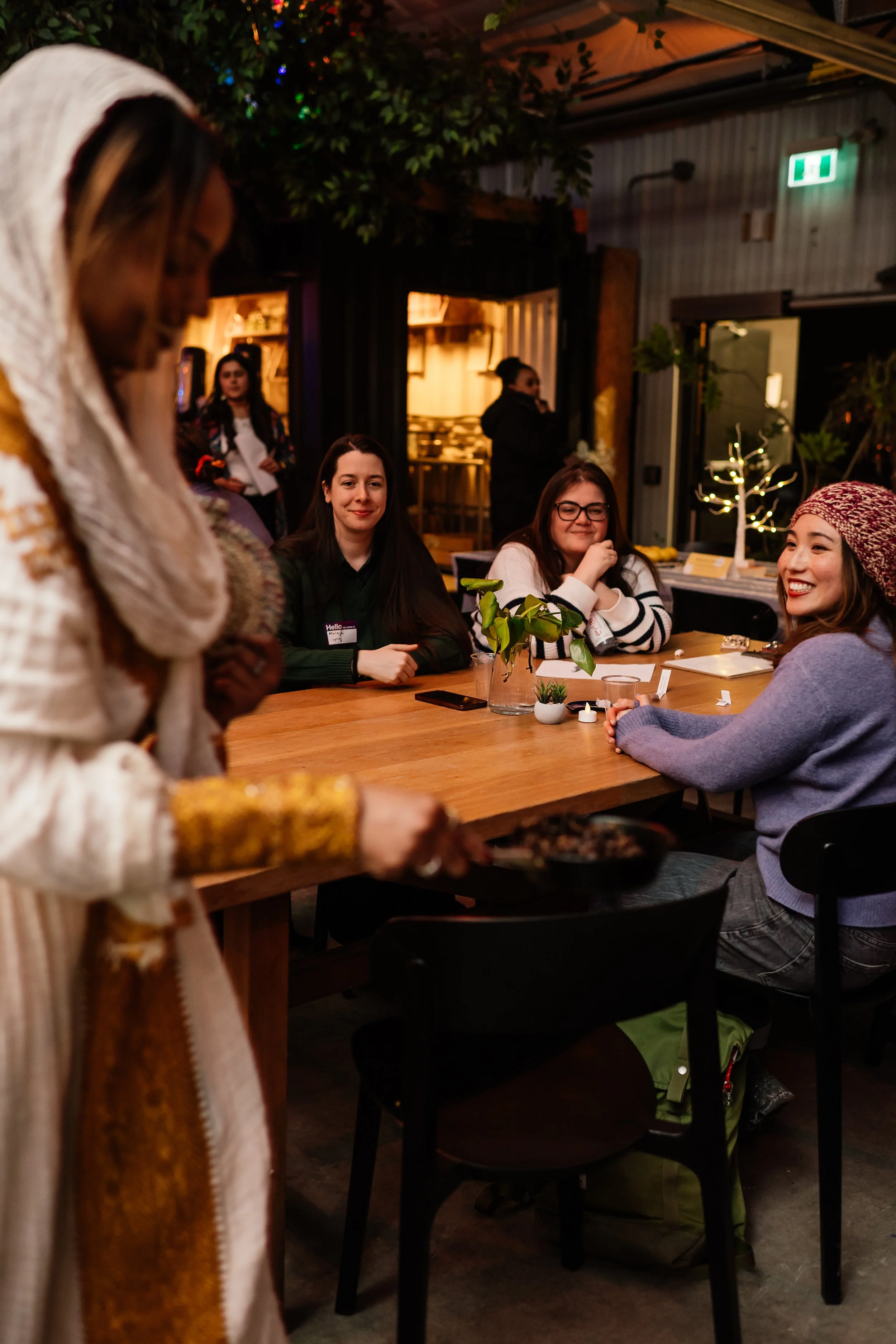 Four women sitting at a wooden table in a cozy indoor space, with a woman serving food in the foreground. The background features warm lighting, small potted plants, and decorative trees, creating a festive and inviting atmosphere.