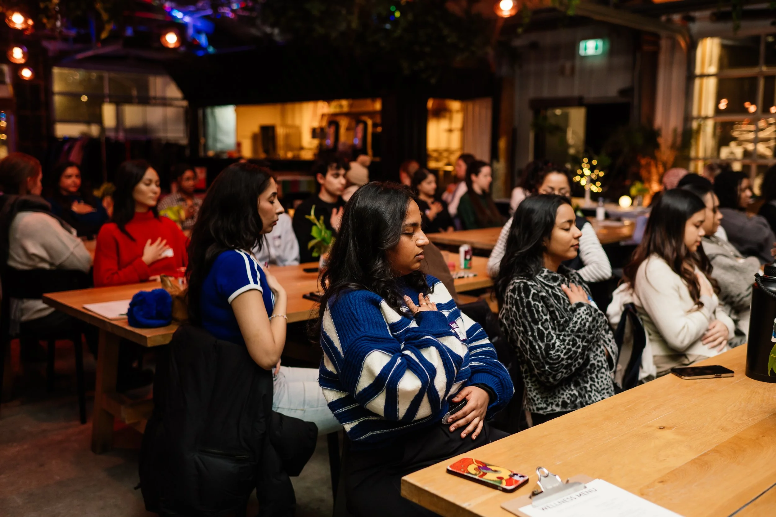 People sitting at tables with their hands over their hearts during a group prayer or moment of silence in a dimly lit indoor event space.
