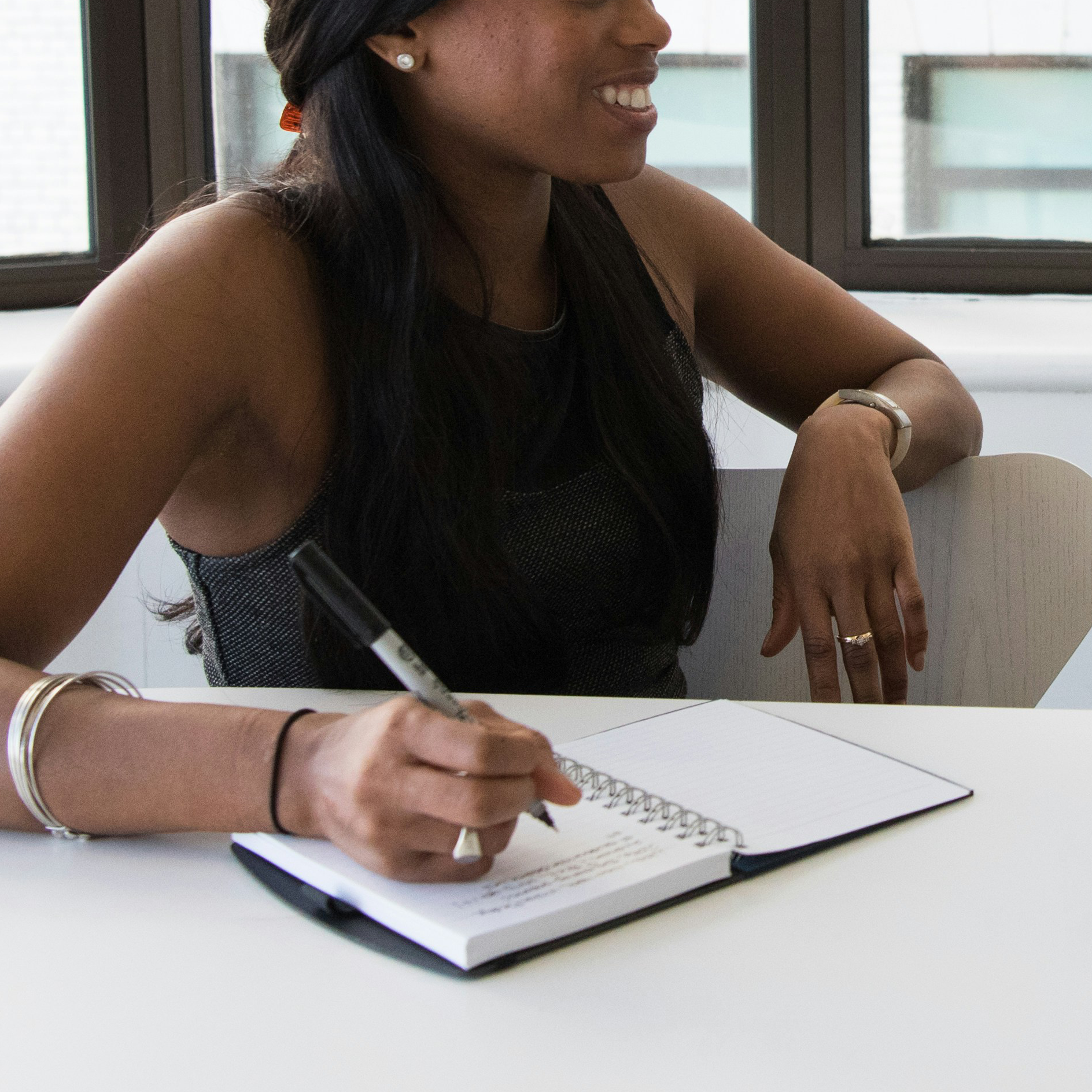 A woman sitting at a white table, smiling, taking notes in a spiral notebook with a black marker, inside a room with large windows.