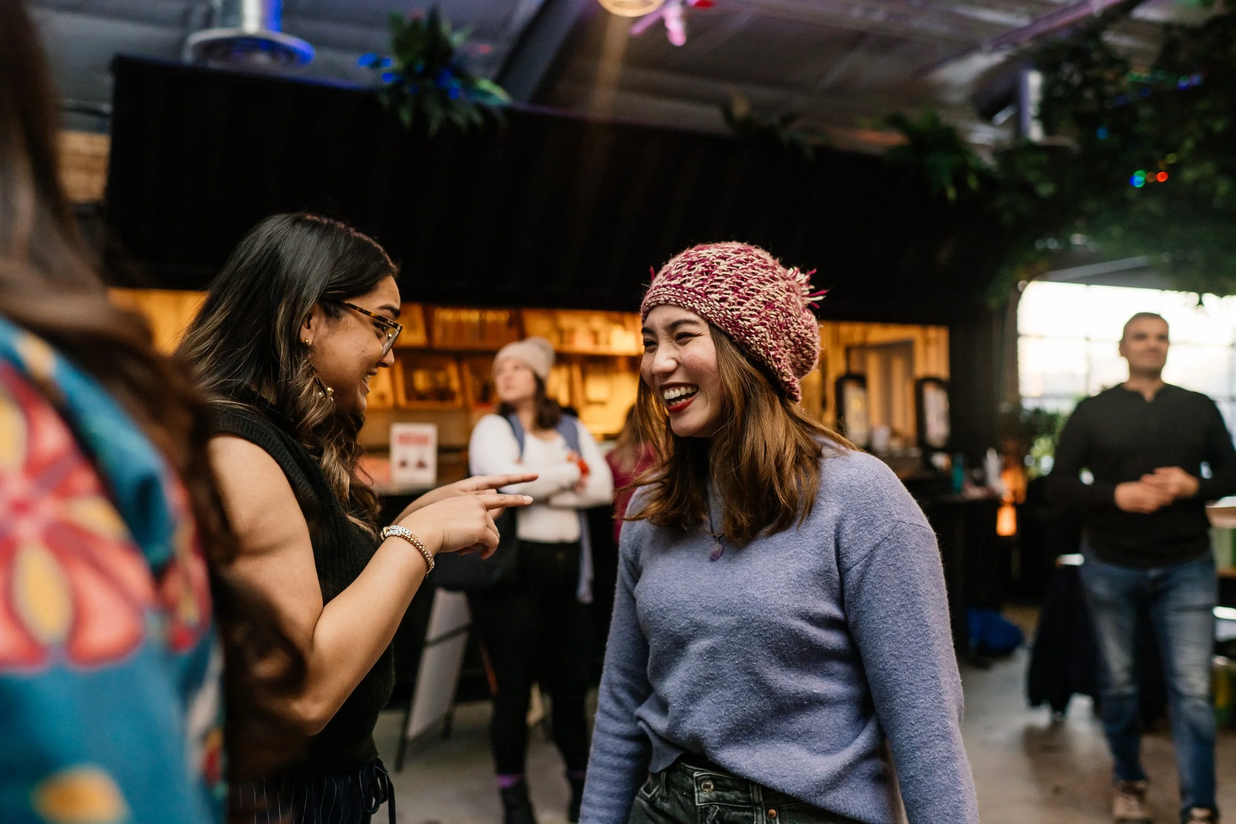 Two women laughing and talking indoors, one woman wearing glasses and a black sleeveless top, the other wearing a pink knit hat and a purple sweater, with other people in the background.
