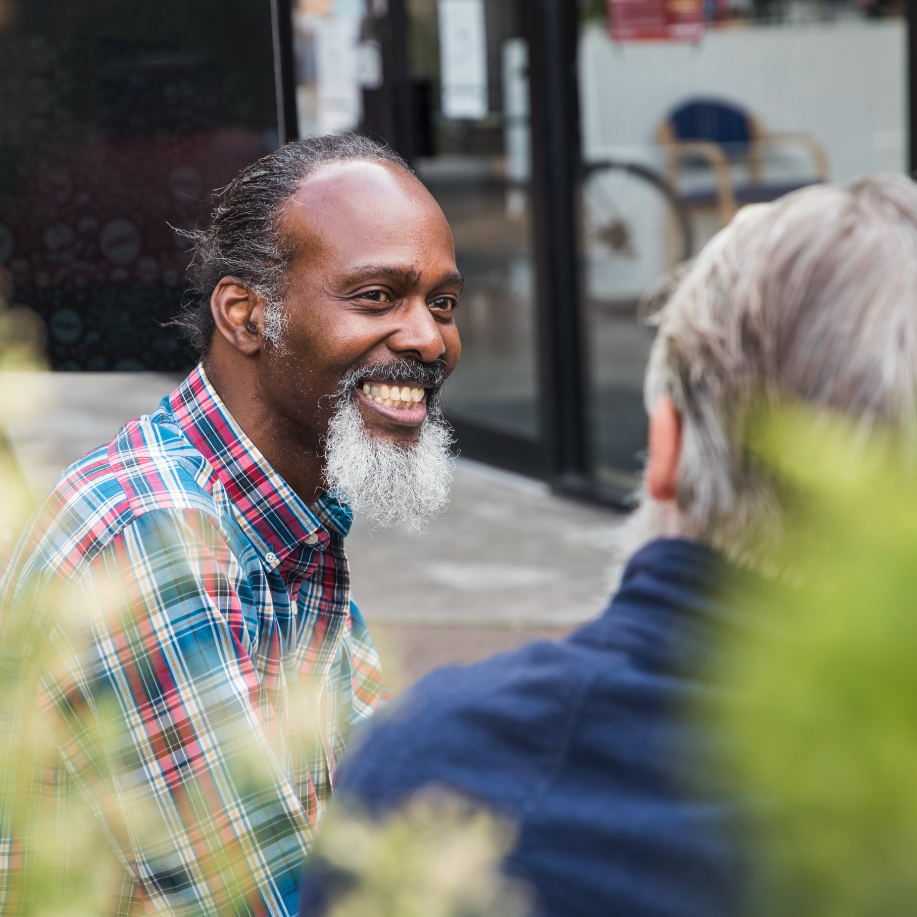 Two men talking outdoors, one smiling in a plaid shirt, the other in a dark jacket, with blurred plants in the foreground.