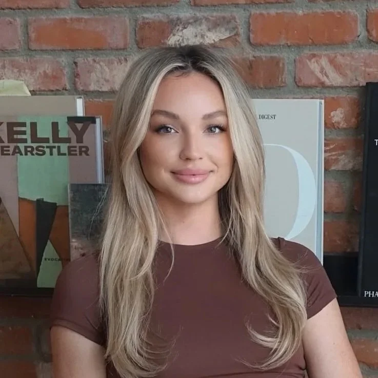 A young woman with blonde hair and a brown shirt posing in front of a brick wall with magazines and posters in the background.