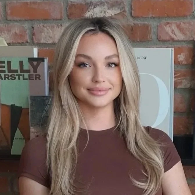 A young woman with blonde hair and a brown shirt posing in front of a brick wall with magazines and posters in the background.