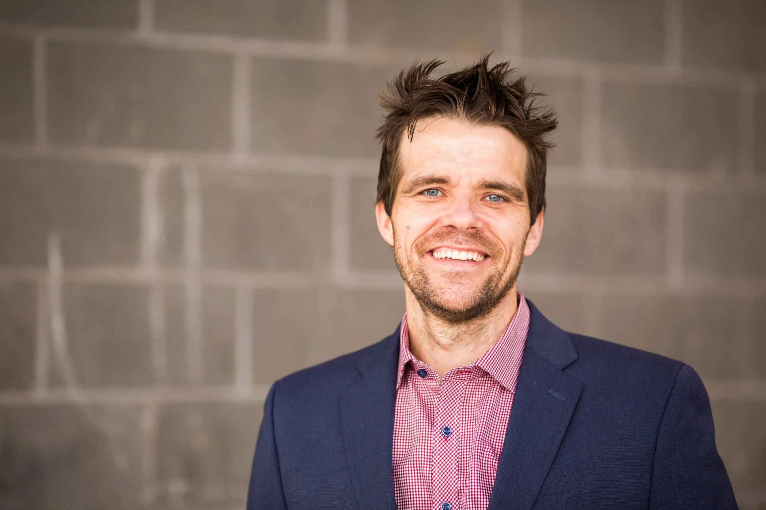 A man smiling, wearing a blue blazer and red checkered shirt, standing against a gray brick wall.