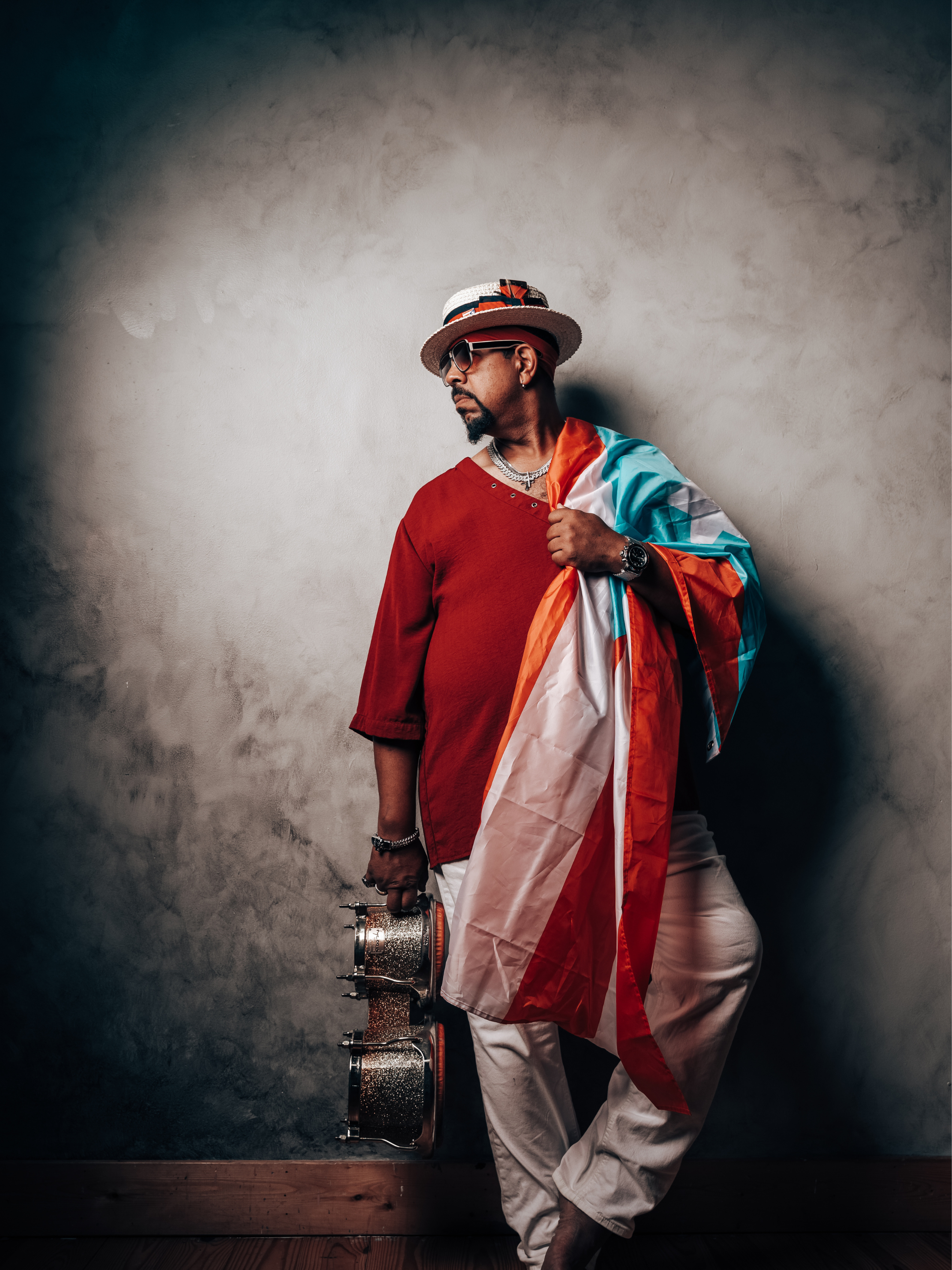 A man dressed in a red shirt, white pants, wearing sunglasses and a straw hat, holding a flag and a drum, standing against a textured wall.