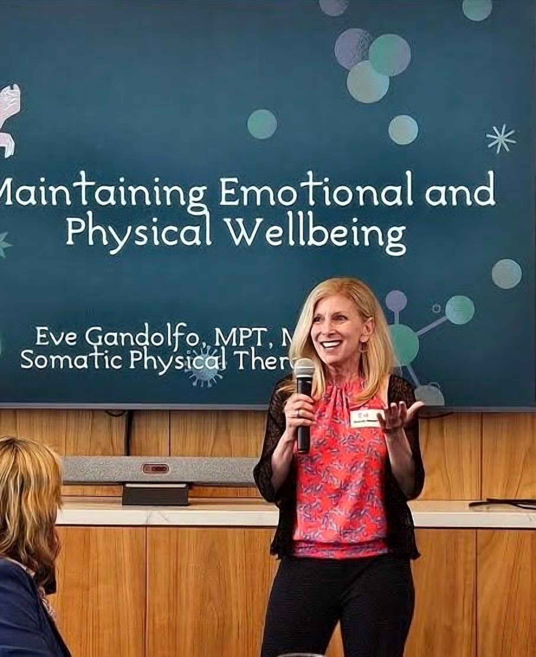 A woman with blonde hair, smiling, speaking into a microphone at a presentation or lecture. She is wearing patterned top. There is a large screen behind her with text about maintaining emotional and physical wellbeing, and part of her face is visible in the image.