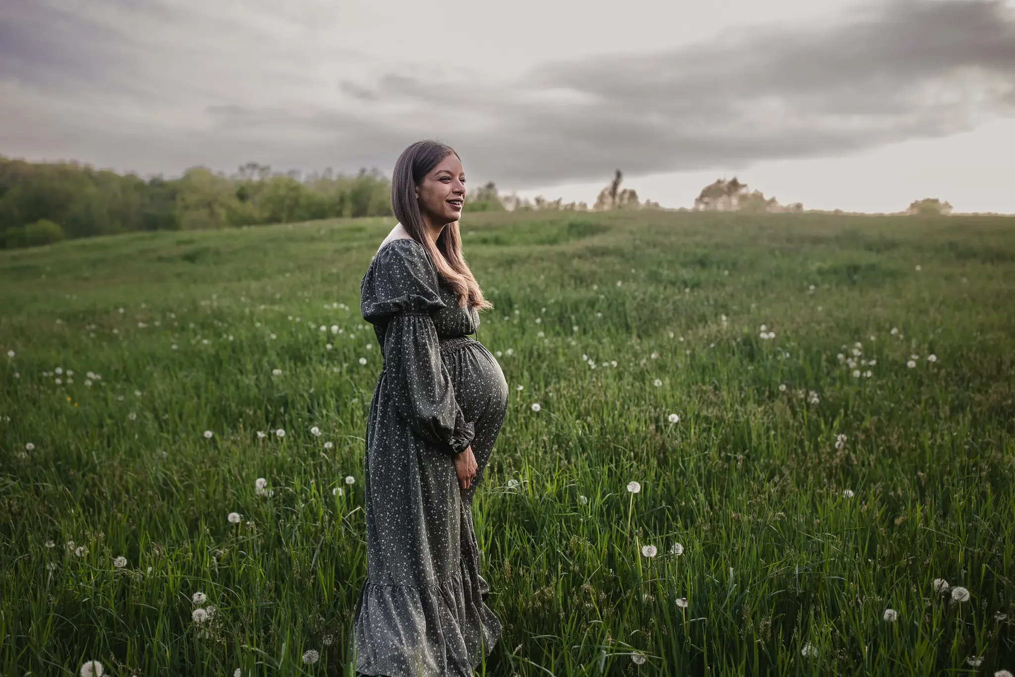 A black pregnant mother holding her belly stands on a grassy hill. Grand Rapids Lifestyle Maternity Photography session by Brianna Trammell of The Nurturing Company.