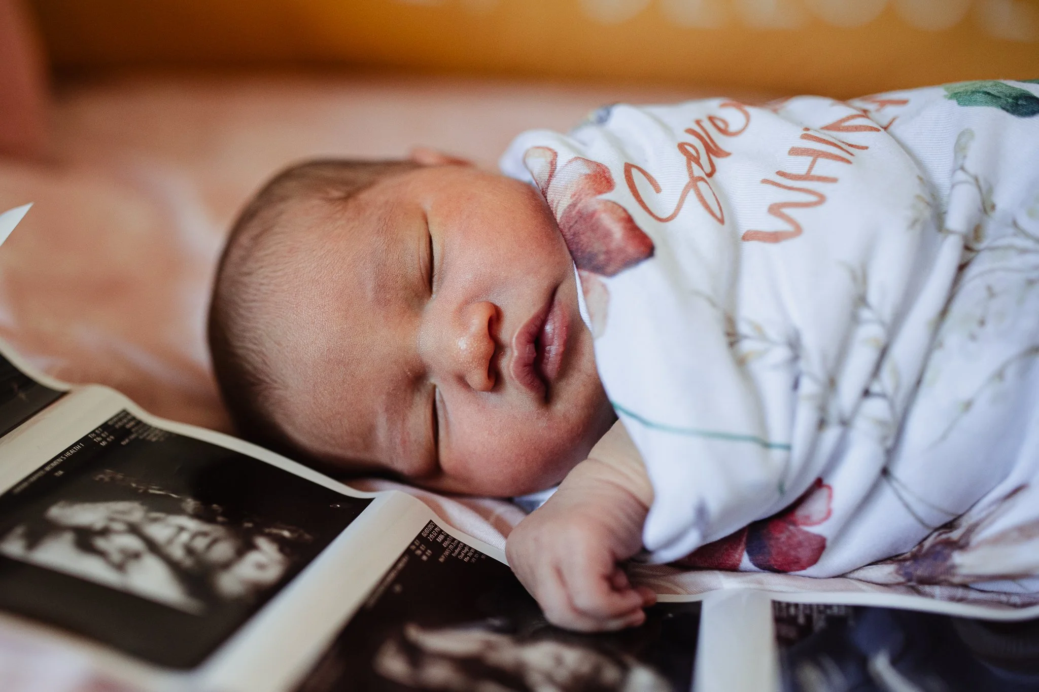 A newborn lies swaddled on a bed with her hand resting on sonogram photos. Grand Rapids Lifestyle Newborn Photography session by Brianna Trammell of The Nurturing Company.