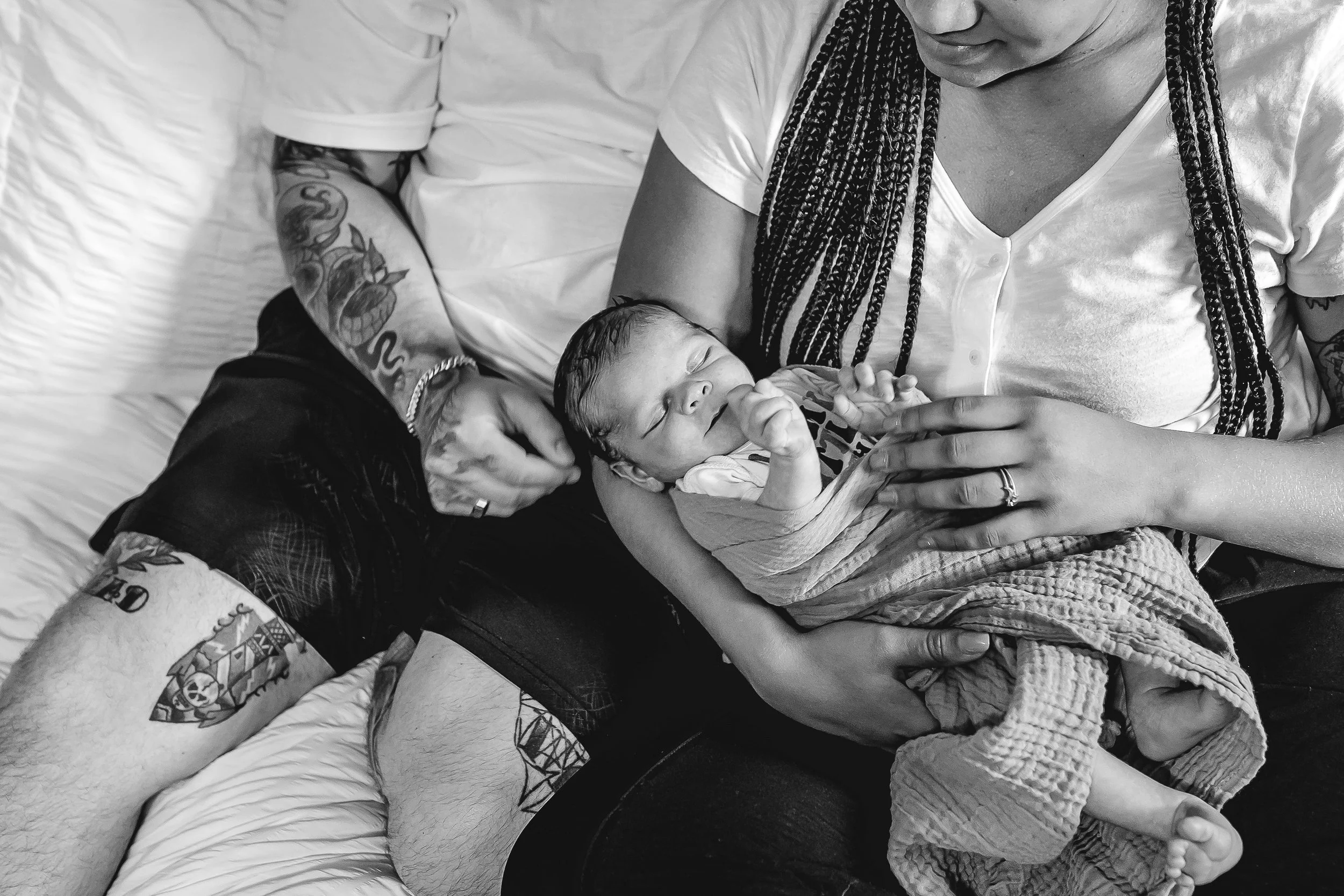A couple sitting closely on a bed gently cradles their sleeping newborn. In-home newborn photo session by Randi Armstrong of The Nurturing Company, a doula and photography business in Grand Rapids, Michigan.