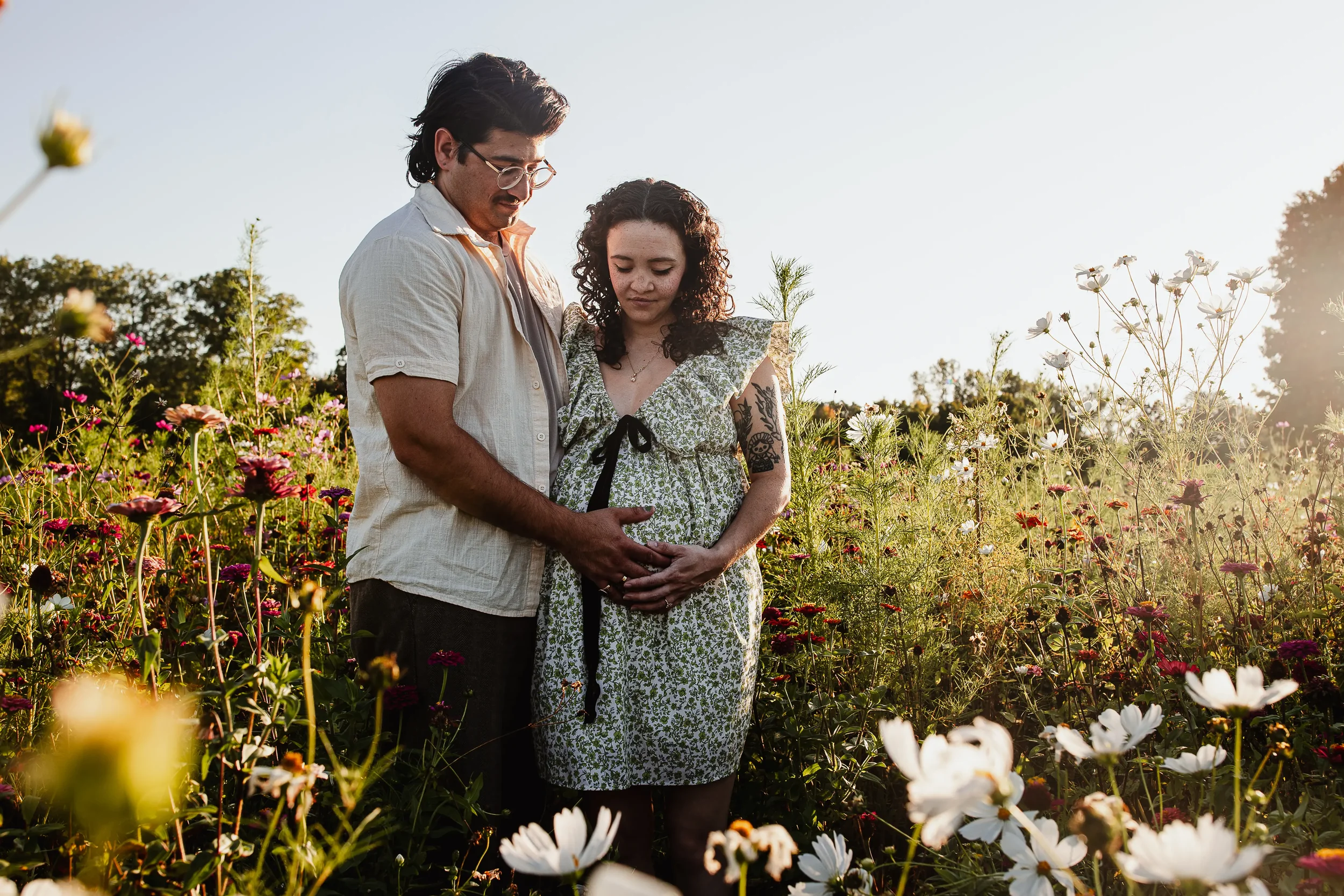 A man and a pregnant woman cradle her pregnant belly while standing in a field full of flowers. Grand Rapids Lifestyle Maternity Photography session by Brianna Trammell of The Nurturing Company.