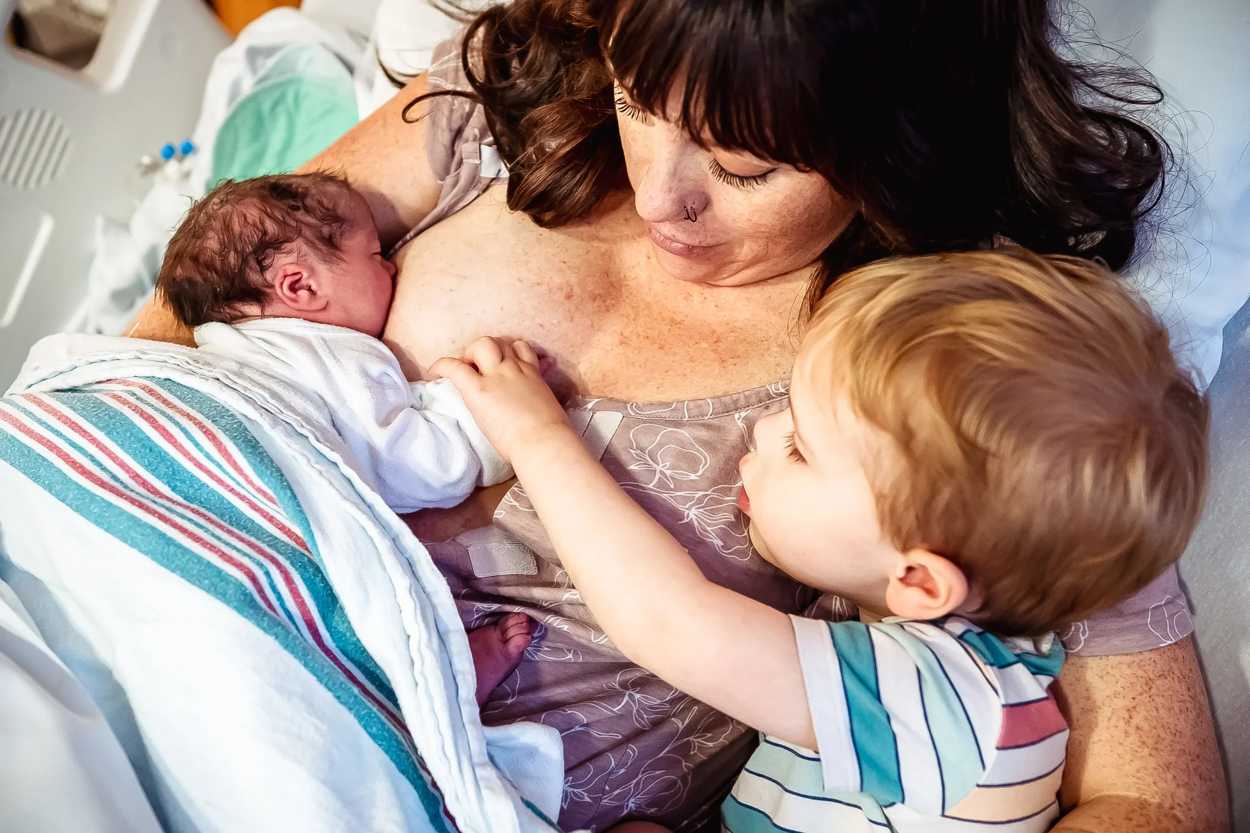 Woman breastfeeds her newborn while her toddler holds the newborn's hand. Big brother meets the newest member of the family.