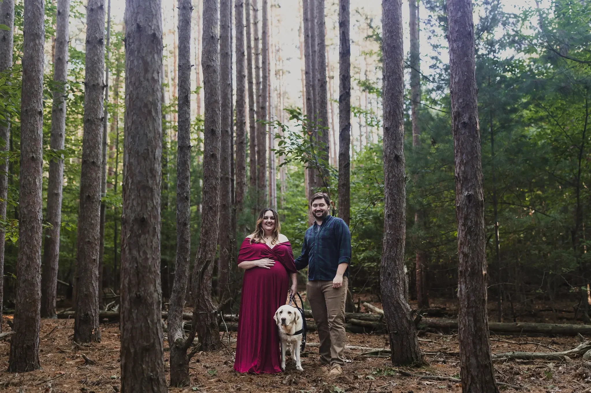 A pregnant mother holding her belly stands next to her partner and support dog in the forest. Grand Rapids Lifestyle Maternity Photography session by Brianna Trammell of The Nurturing Company.