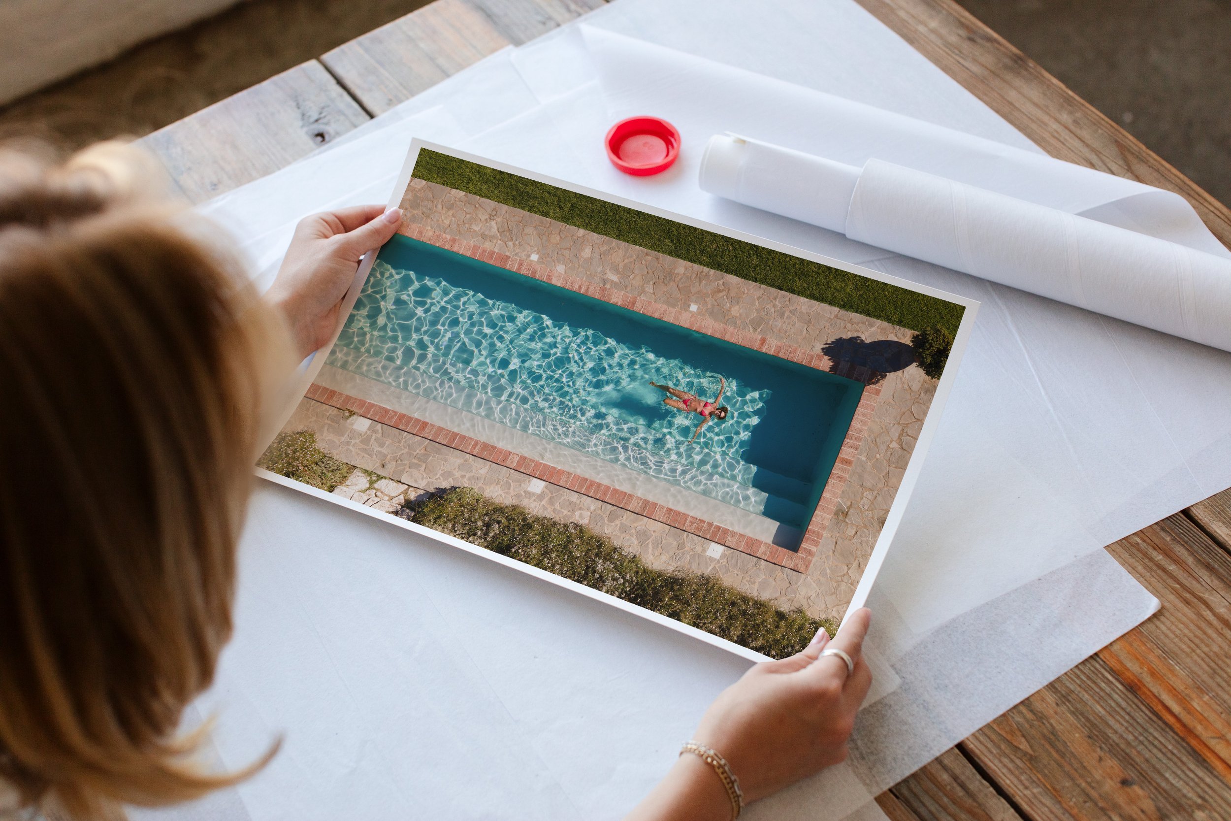 Person holding a printed photo of a swimming pool with a child swimming in it, on a white table with rolled-up papers and a red bottle cap.