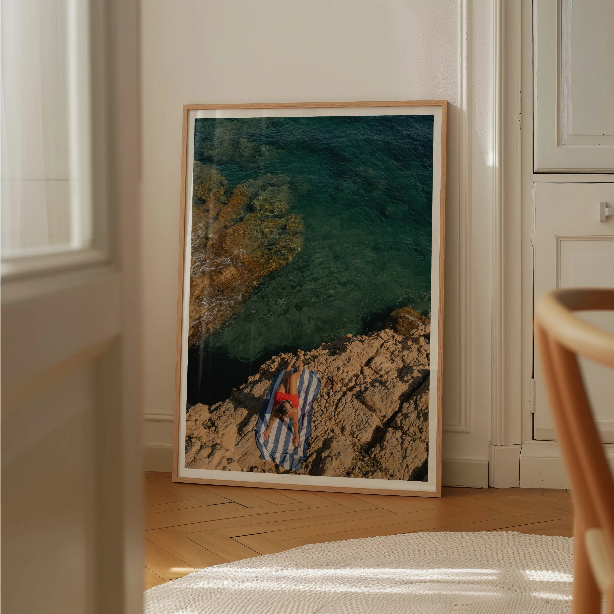 A framed photograph of a rocky coastline and ocean leaning against a white wall in a room with light wood flooring.