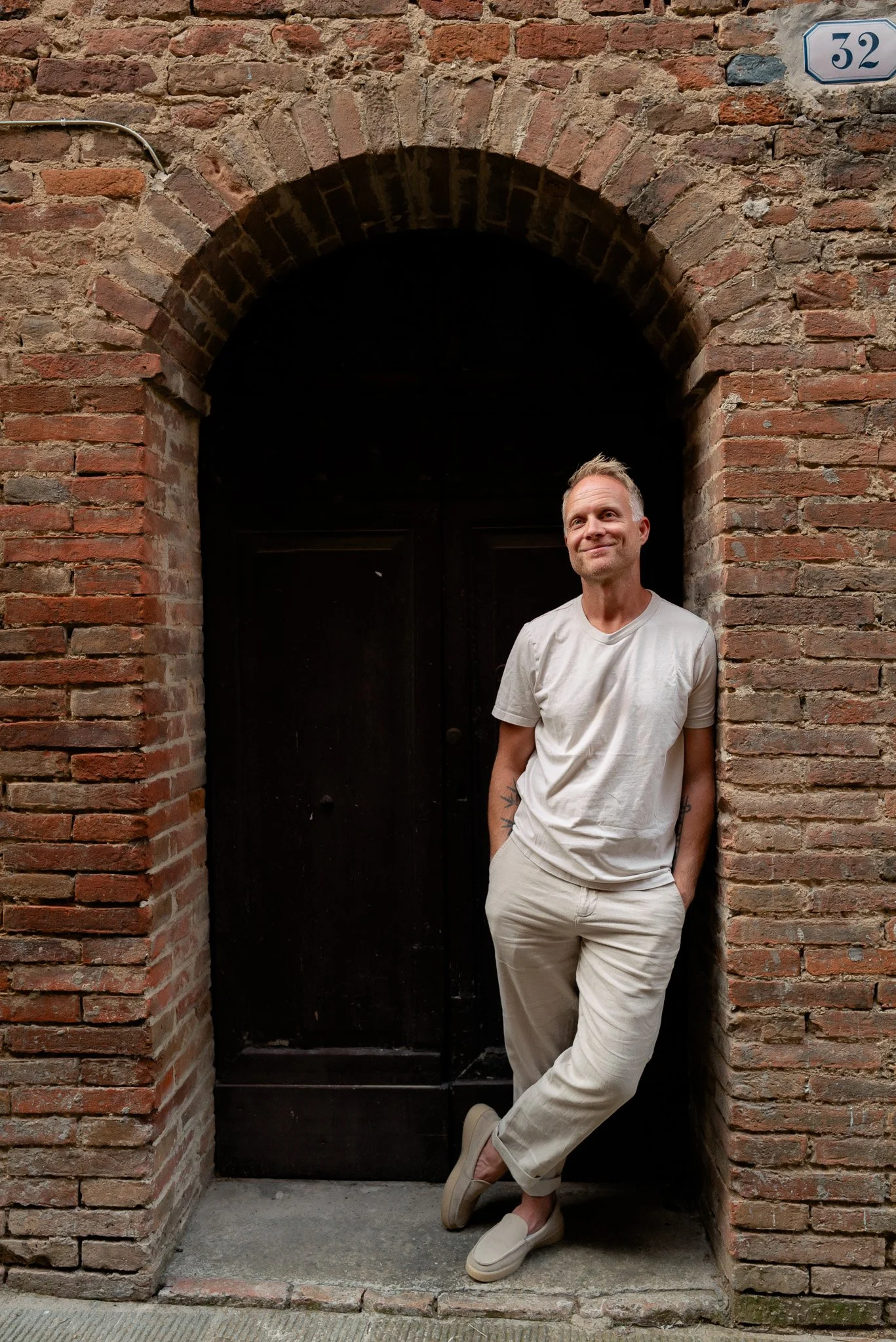 A man wearing a light-colored t-shirt and pants standing against a brick wall with an arched doorway, smiling with one hand in his pocket, in front of a brick and brick-lined doorway.