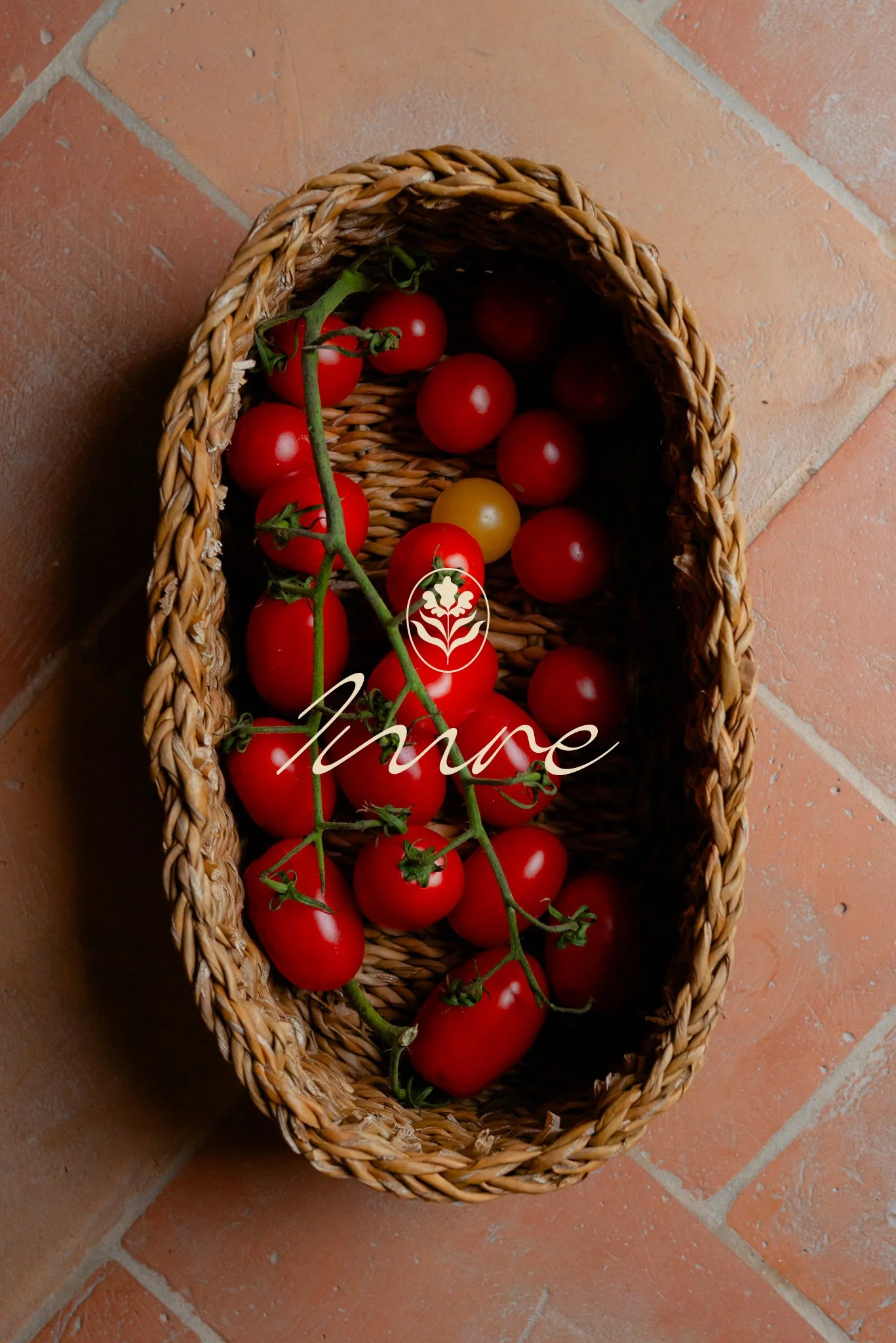 A woven basket filled with ripe cherry tomatoes, some still attached to green vines, placed on a terracotta tiled floor.