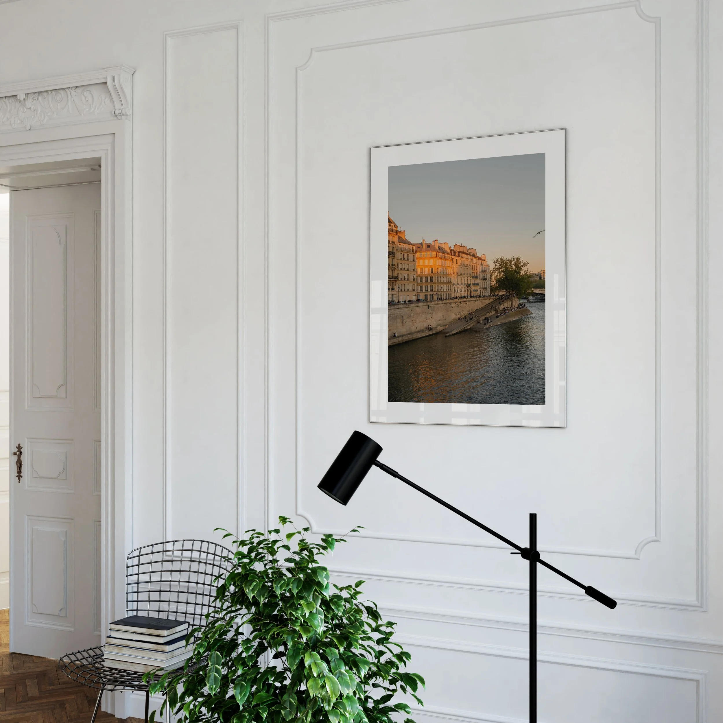 Interior view of a room with white paneled walls, a large mirror showing a cityscape with traditional European-style buildings along a river, a black adjustable floor lamp, a wire chair with a stack of books and a plant underneath, and a frame on the