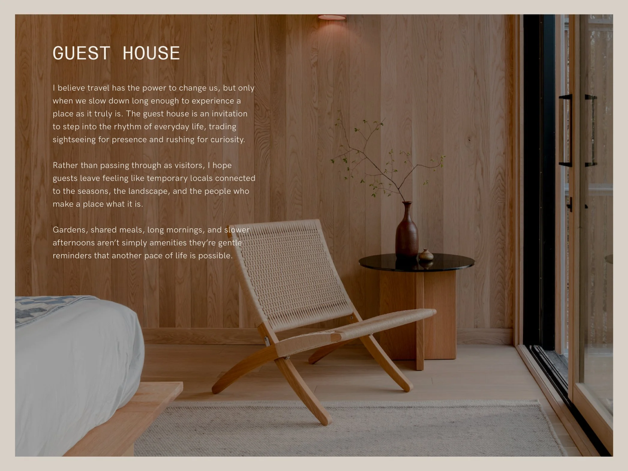 Interior of a guest house room with wooden wall paneling, a beige woven chair, a small round black table with a brown vase and a branch with green leaves, and sliding glass doors leading outside.