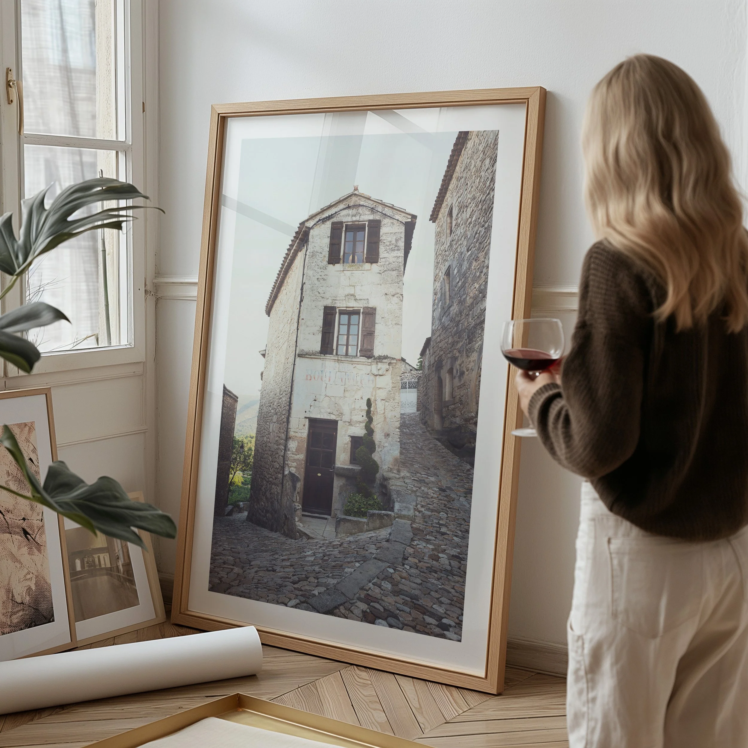 A woman with long blond hair holding a glass of red wine, looking at framed photographs of old buildings in a well-lit room with a window.