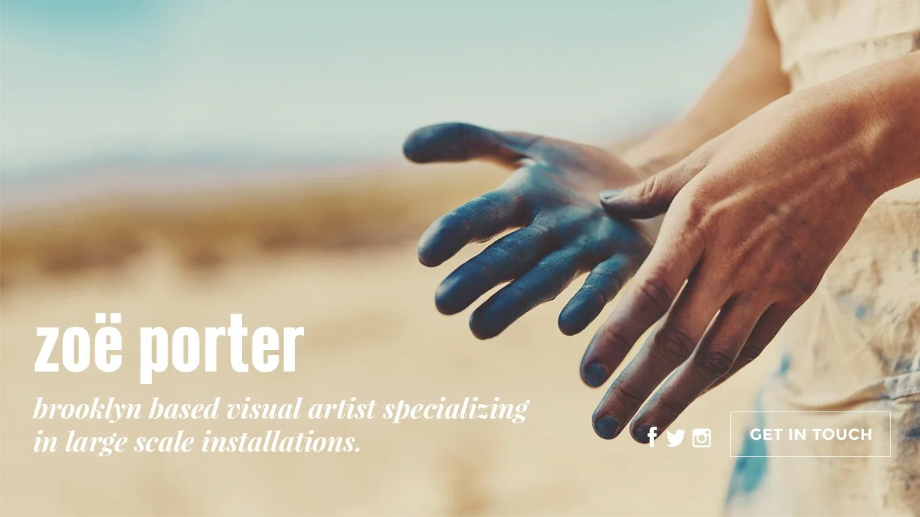 Close-up of a person with paint-stained hands, standing outdoors on a beach or desert landscape, with the person's hands stretched out to the side.