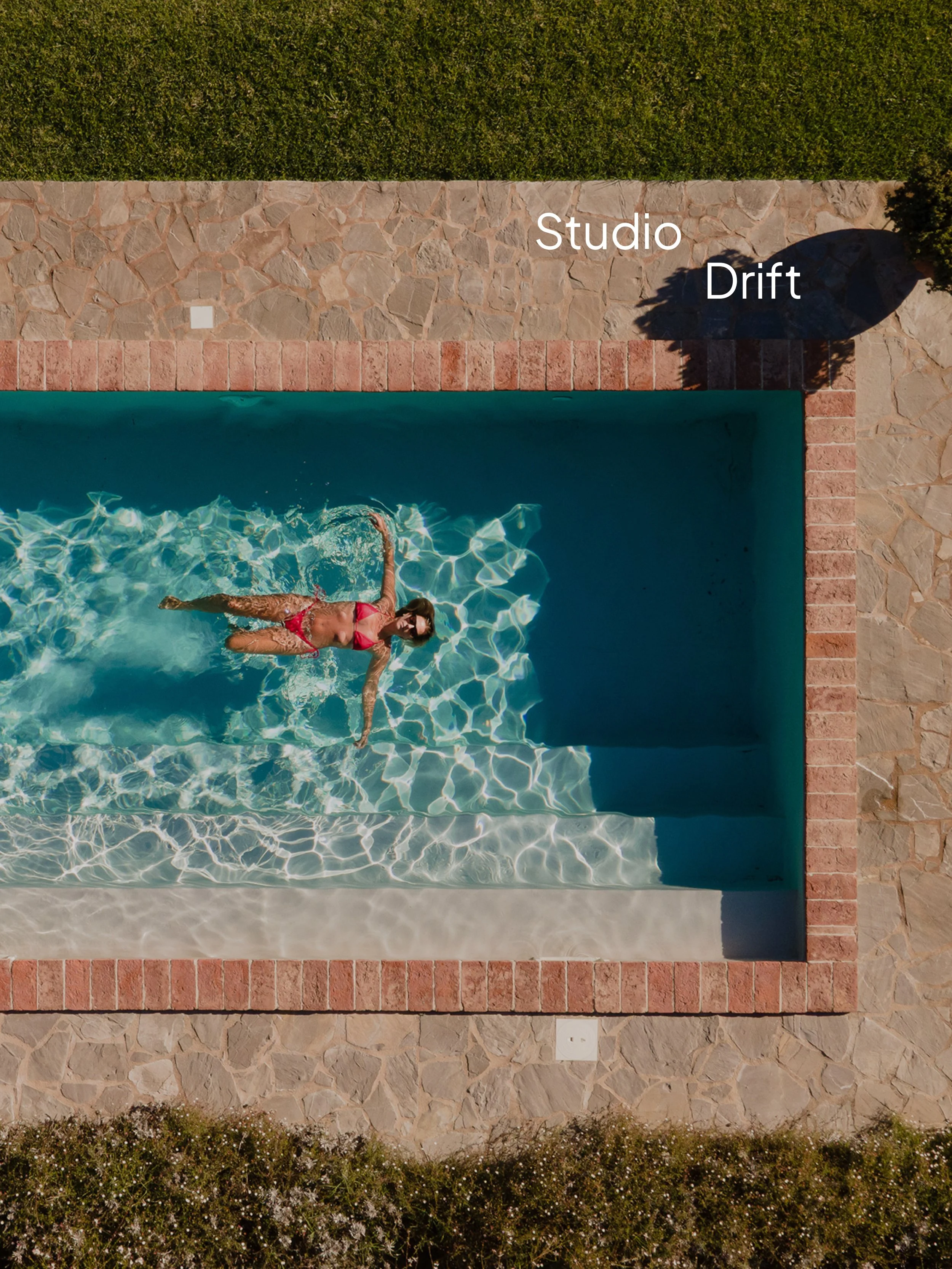 A woman in a pink swimsuit floating and relaxing in a swimming pool, surrounded by a stone and brick border with green bushes and grass nearby.