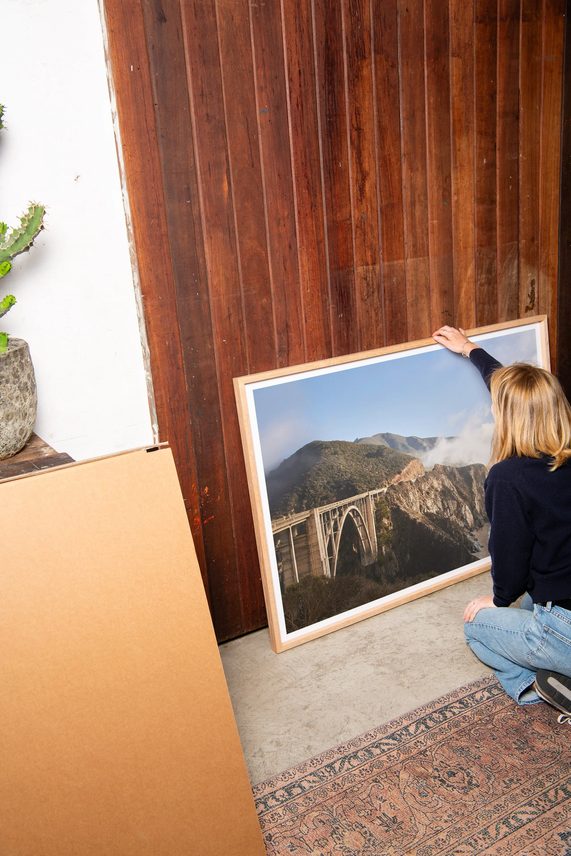 Woman in black sweater and jeans kneeling on Persian rug, holding a large framed landscape photograph of mountains and a bridge, in a room with wooden wall, cactus in pot, and blank cardboard panel.