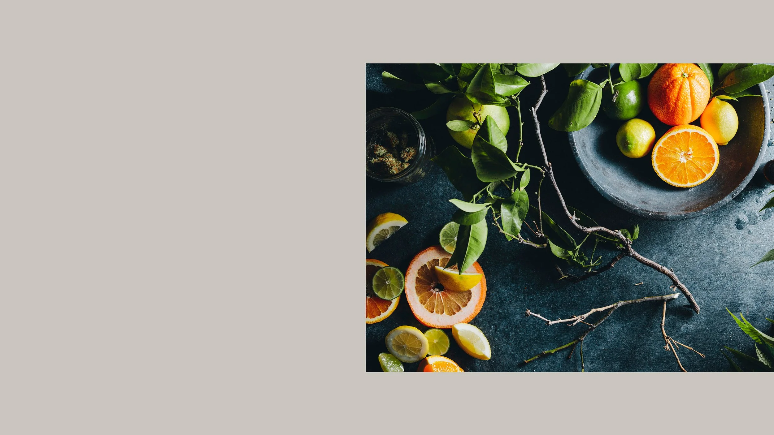 Sliced citrus fruits including oranges, lemons, and limes, along with whole oranges and citrus on a dark surface with green leaves and branches.