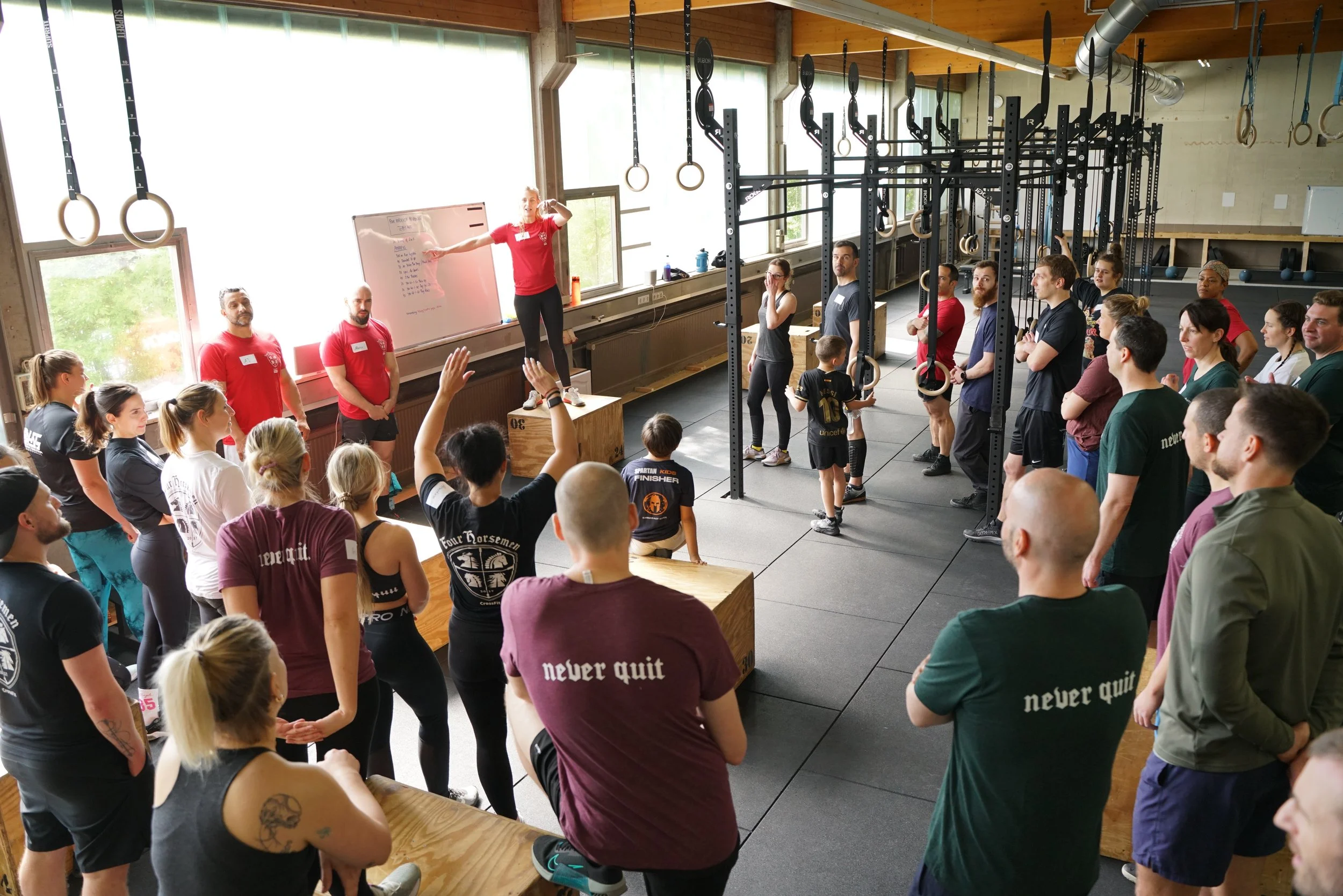 Menschen in einem Fitnessraum während eines Workshops oder Seminars, mit einer Person an einer Tafel, die etwas erklärt. Es gibt Kreißstangen und Gymnastikring im Raum.