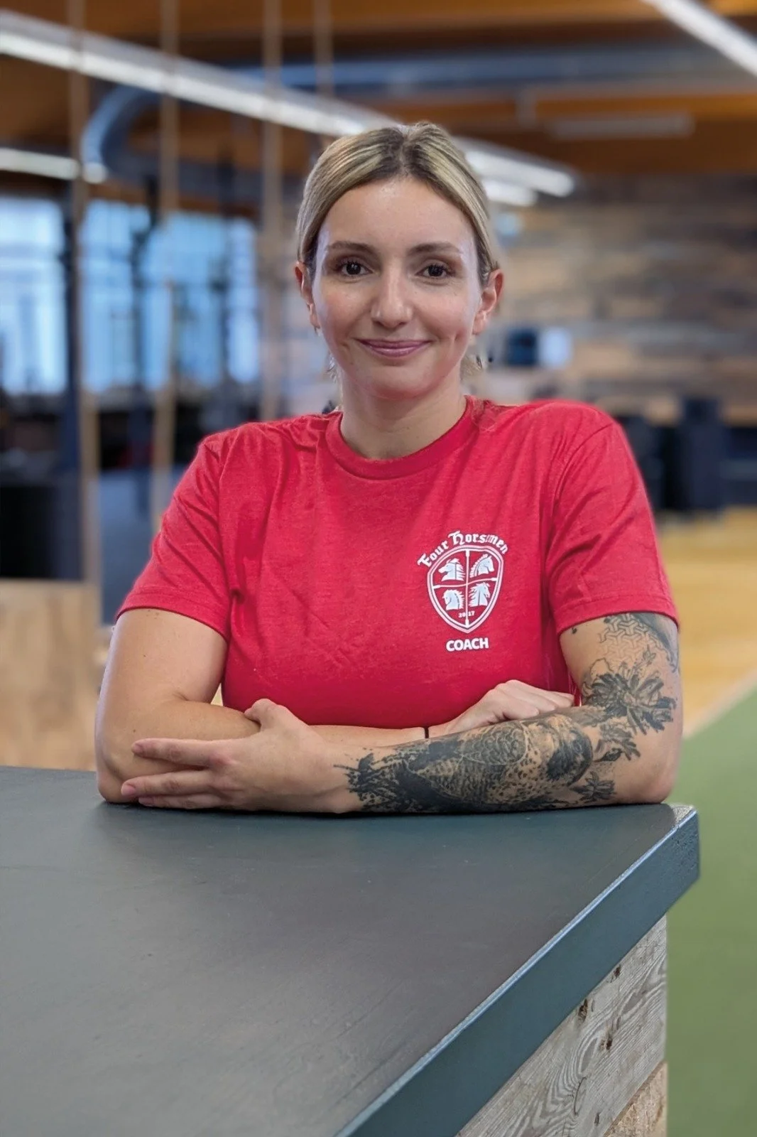 Eine lächelnde Frau mit Tattoos auf den Armen, trägt ein rotes T-Shirt mit einem Logo und der Aufschrift 'Four Horsmen Coach', sitzt an einem Schreibtisch in einer modernen, offenen Raum mit Holzbalken und großen Fenstern im Hintergrund.