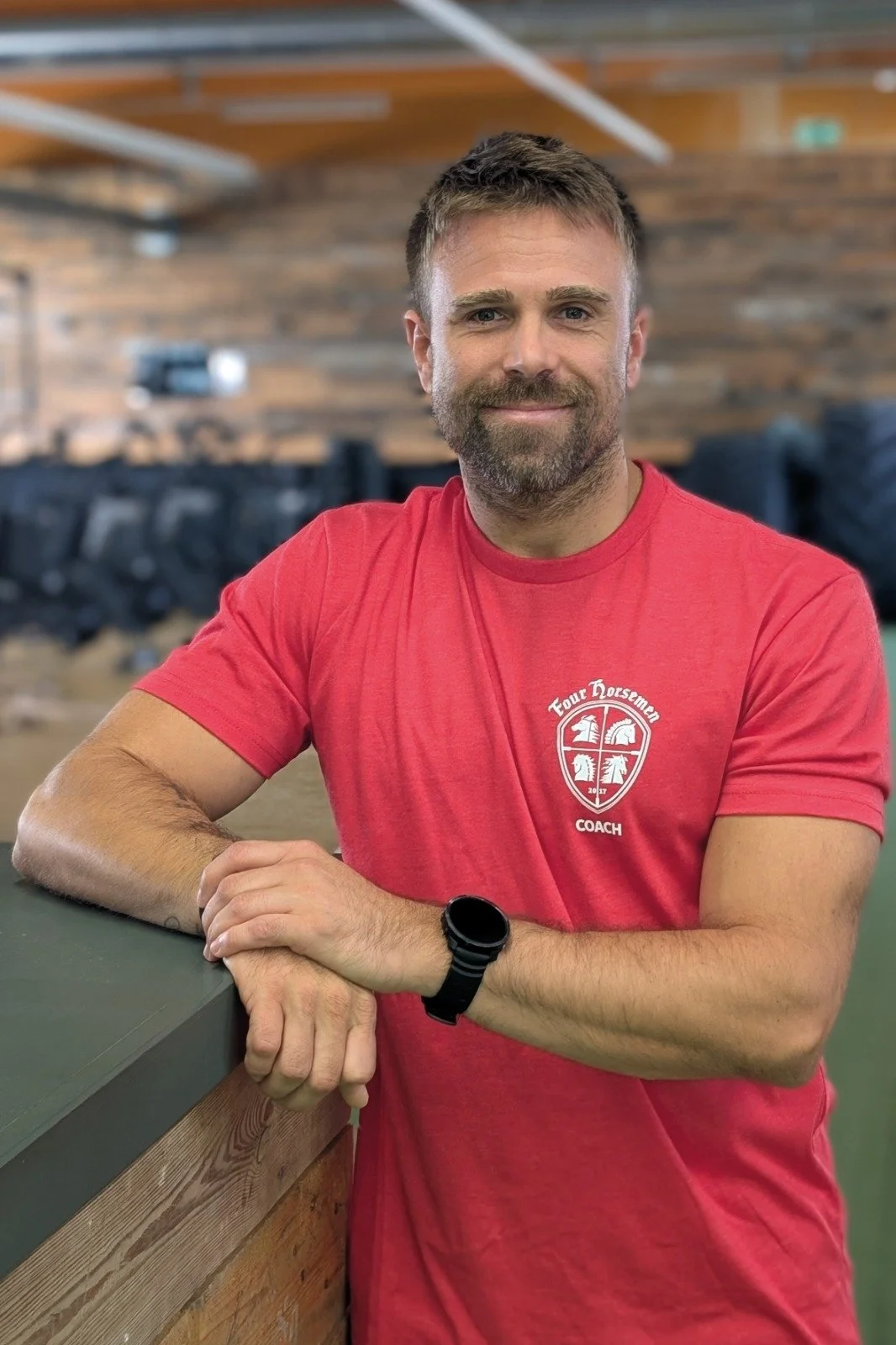 Ein Mann mit braunem Bart und短haar trägt ein rotes T-Shirt mit einem Logo, das 'Tour Horsemen Coach' sagt, und steht in einem Raum mit Holzwänden, lehnt an eine Theke und lächelt.