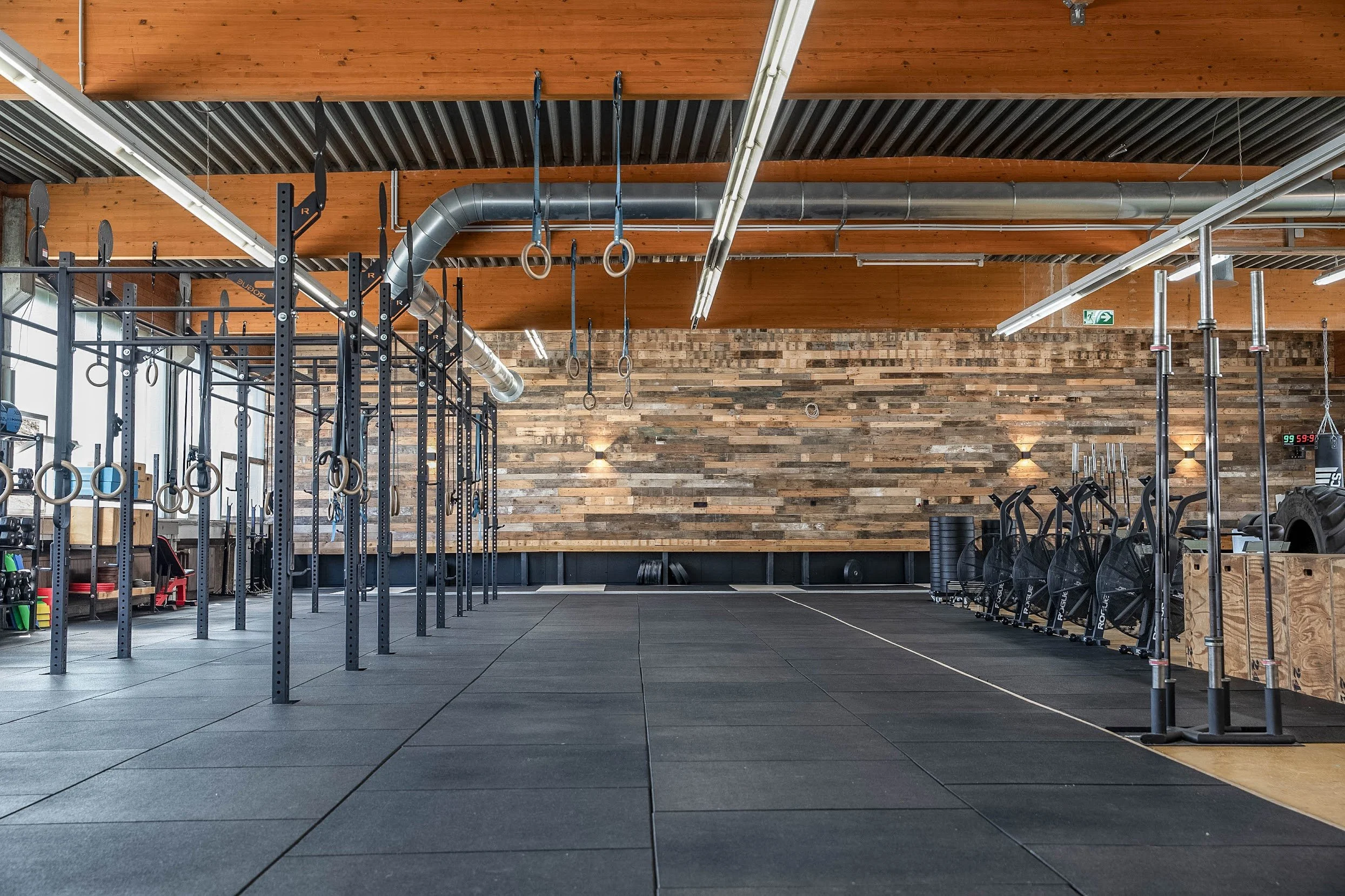 Innenansicht eines Fitnessstudios mit Kraftgeräten, Hanteln, Turnringen und anderen Trainingsgeräten, Holzboden und Wandverkleidung aus Holz.