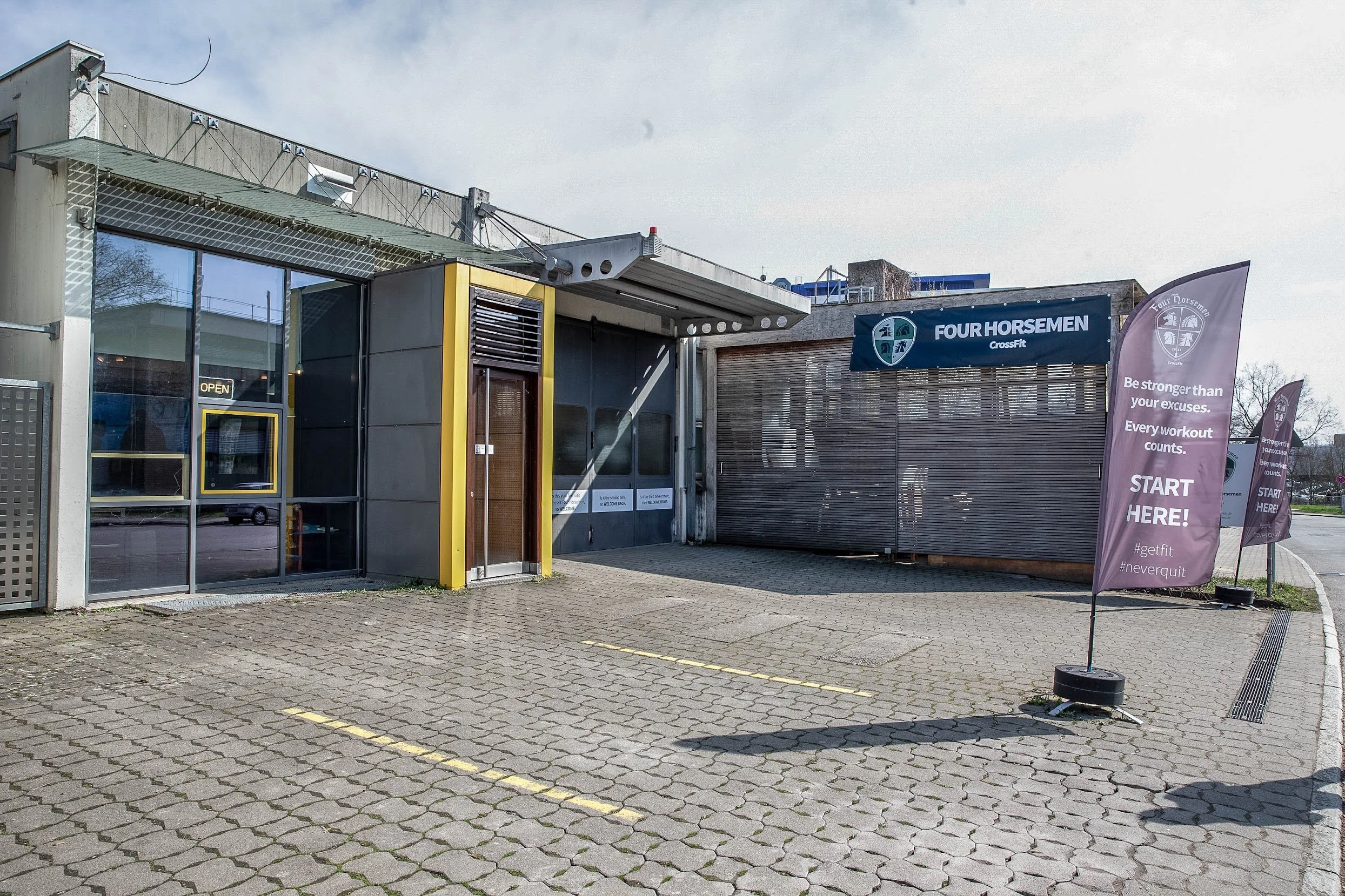 Außenansicht eines Fitnessstudios mit einem blauen Banner, das 'FOUR HORSEMEN CrossFit' zeigt, und pinkfarbene Fahnen mit dem Text 'Start Here!' bei sonnigem Himmel.