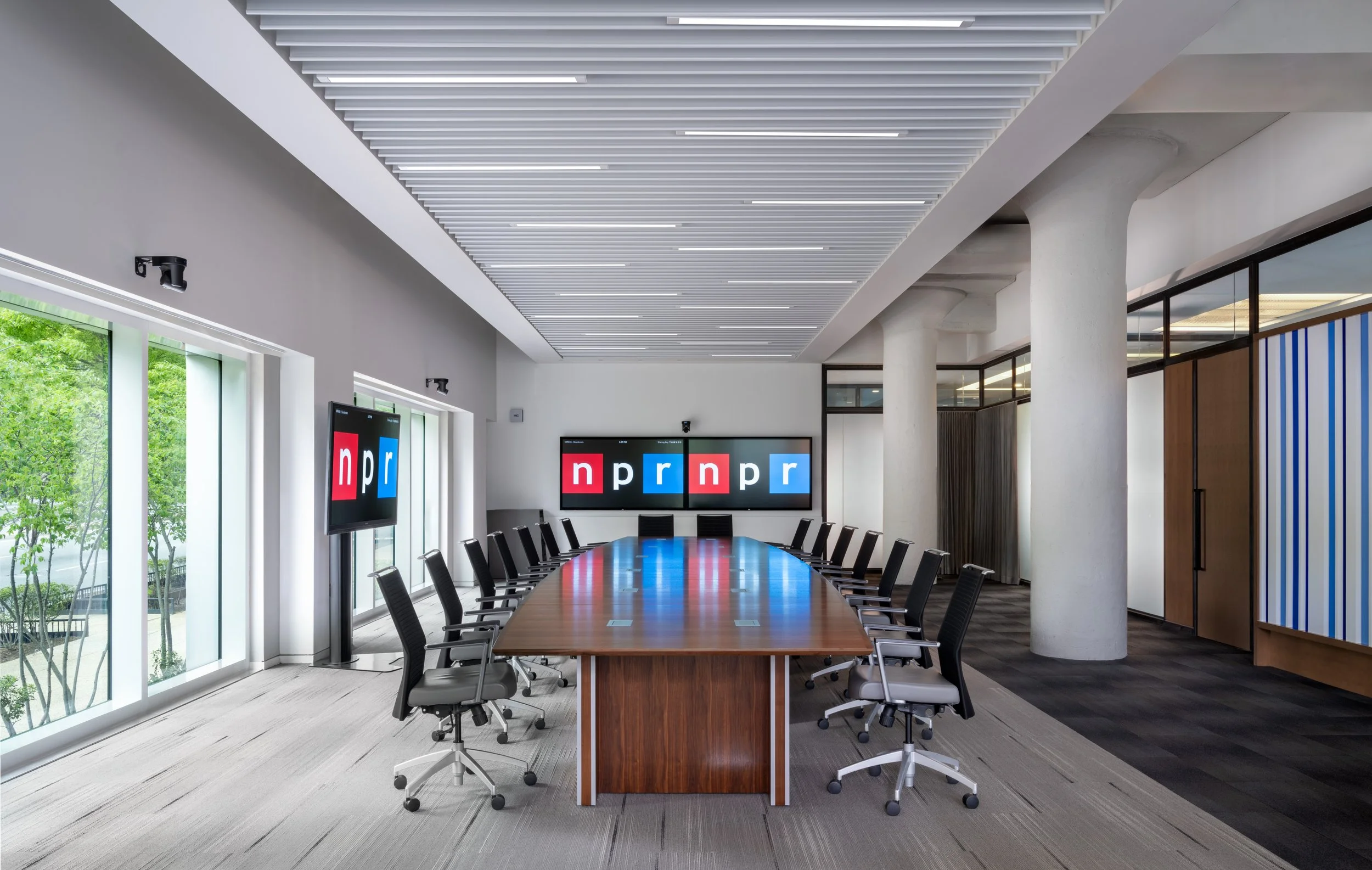 Modern conference room featuring a long wood table surrounded by black task chairs. Three large monitors. The room has full-length windows, white columns, and a slatted ceiling