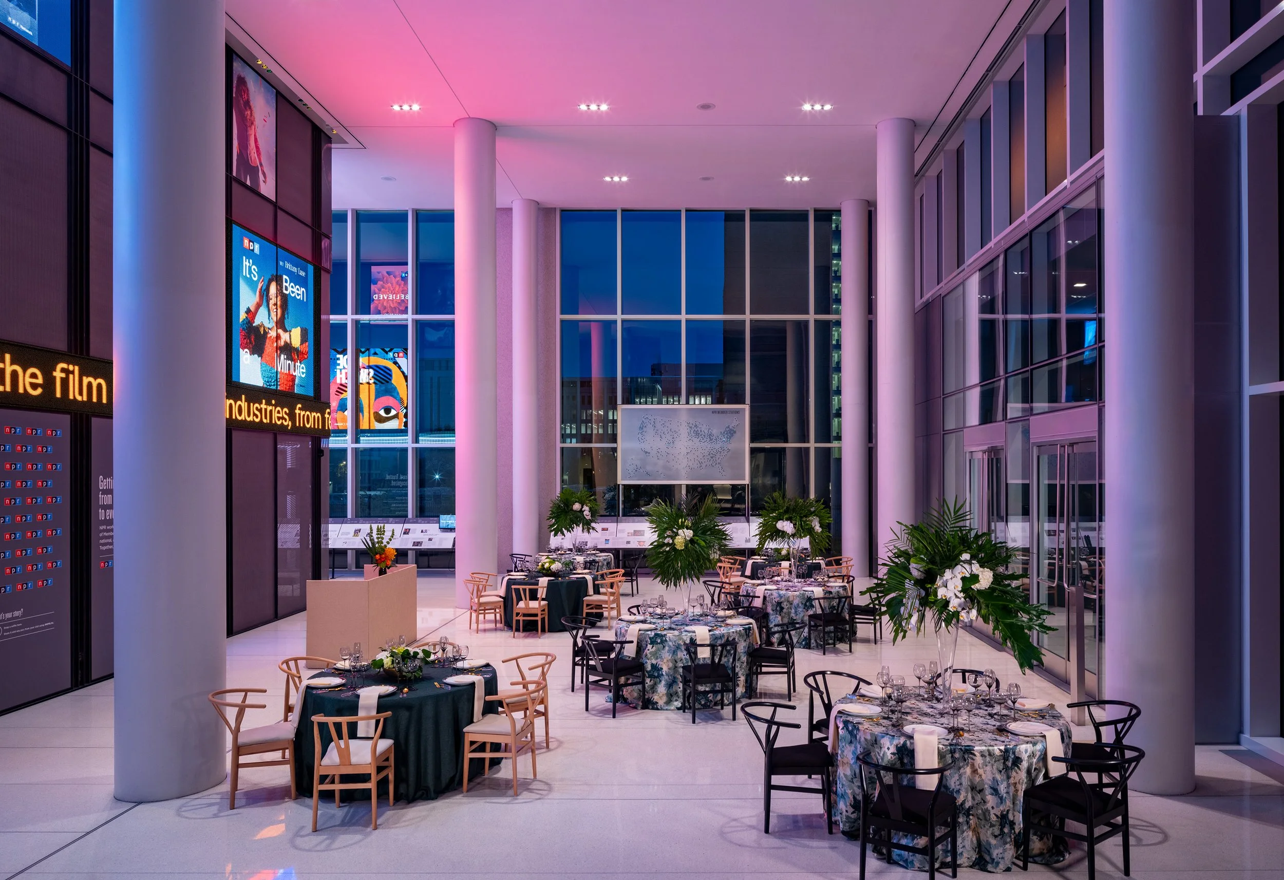 High-ceilinged, modern lobby arranged for dinner, featuring round tables with floral linens, prominent white columns, and large screens with colorful promotional graphics.