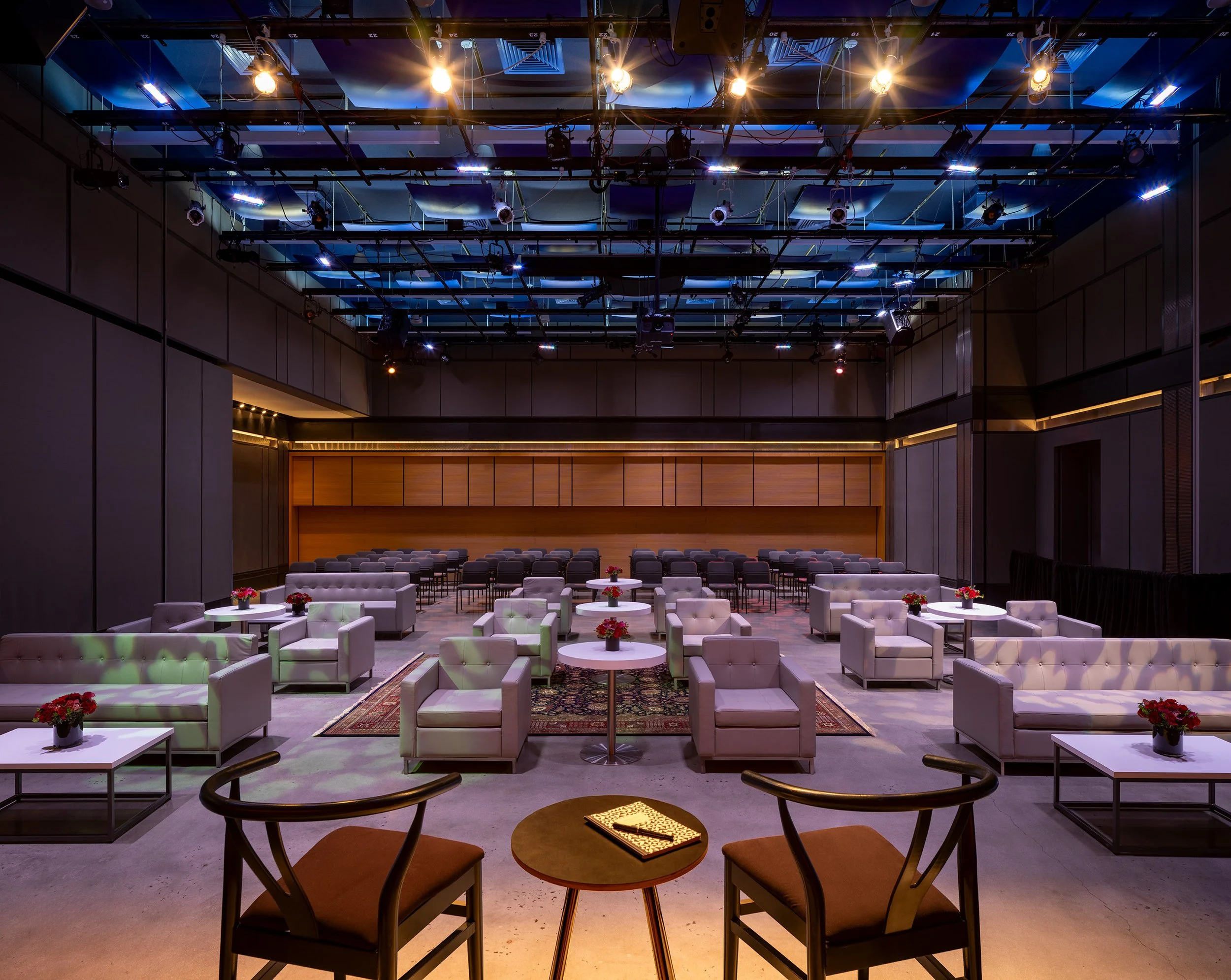  A dimly lit, modern event space with light gray couches and armchairs arranged in conversational clusters around small tables, under a high ceiling with exposed lighting.