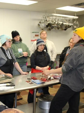 A group of women gathered around a kitchen table, listening to a woman in a yellow beanie and gray shirt demonstrate cooking or food preparation techniques.