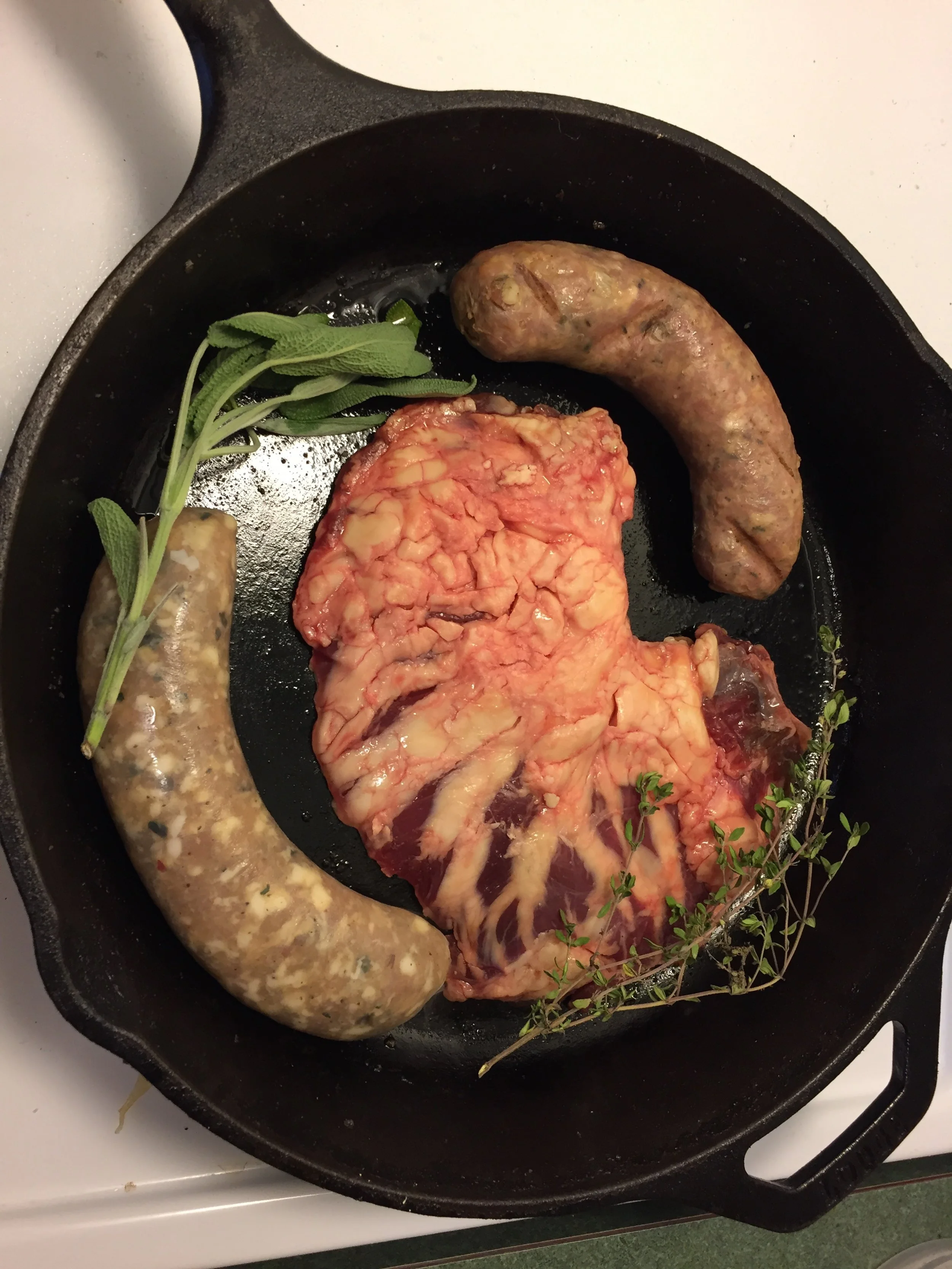 Raw butchered meat, two sausages, and fresh herbs in a frying pan.