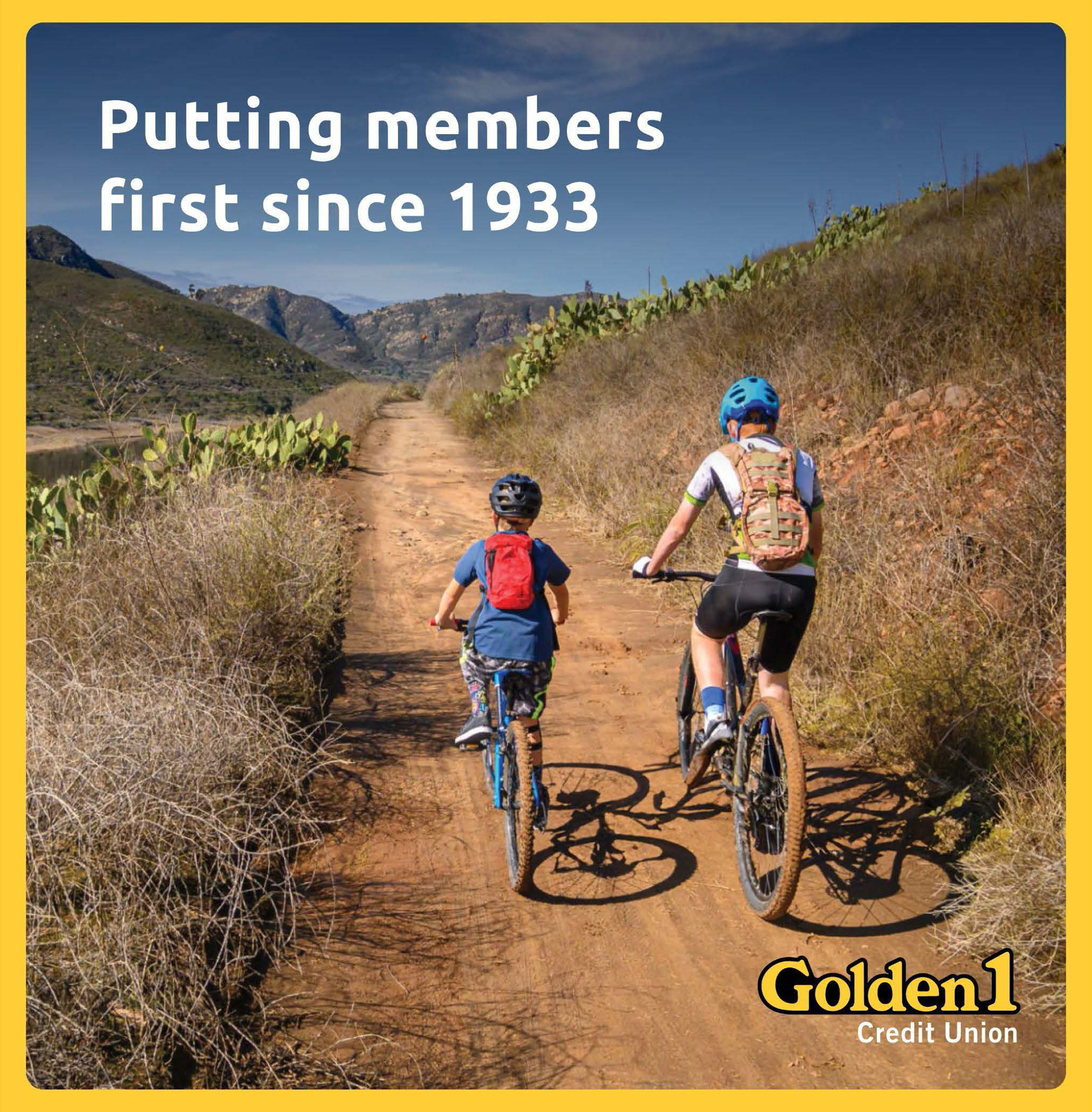 Two children riding bicycles on a dirt trail in a rural, hilly landscape with cacti and dry shrubs, under a blue sky with some clouds.