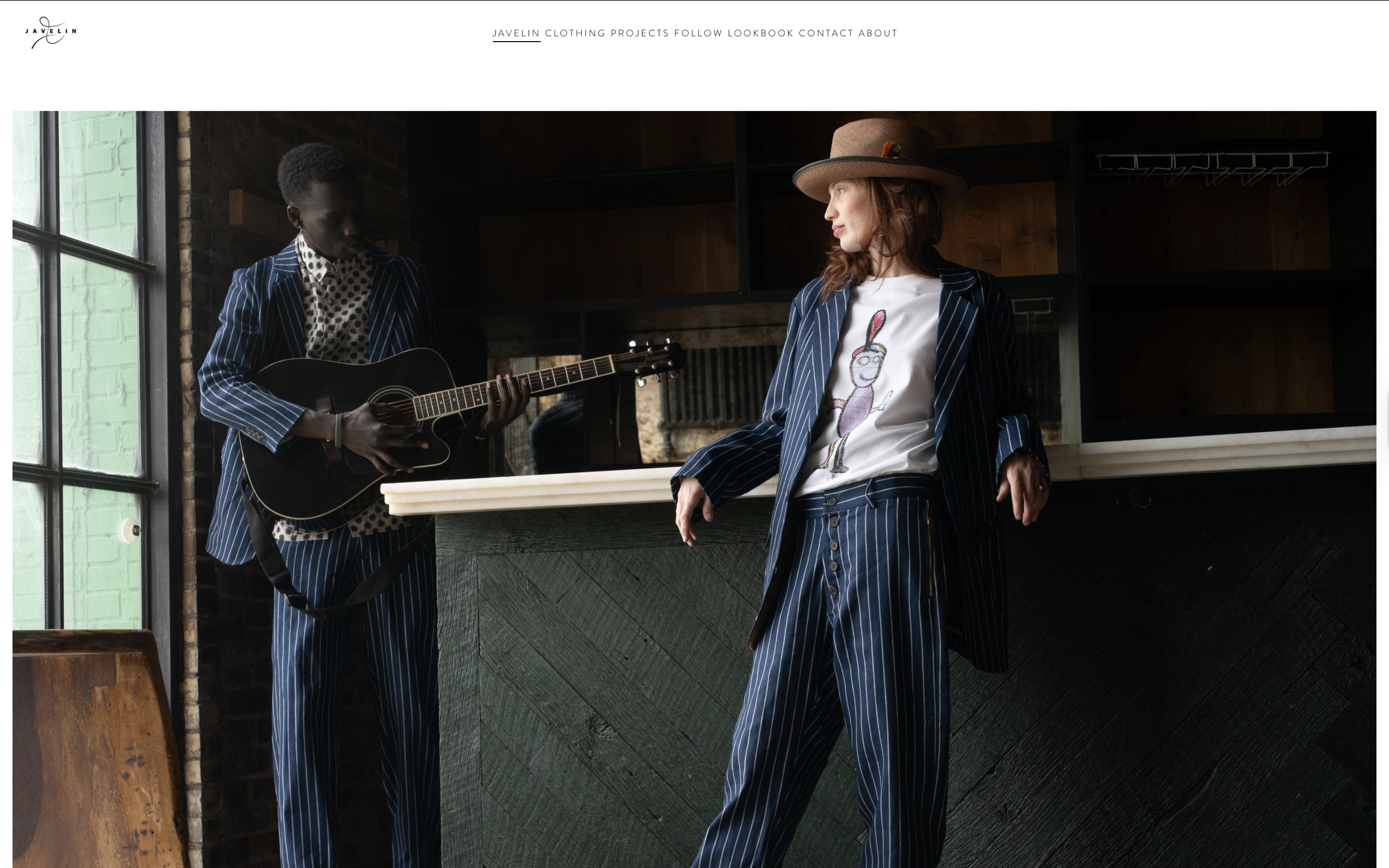 Two women with striped suits and hats in a rustic, loft-style room. One is playing guitar, the other is about to sing. Natural light from large windows illuminates the scene.