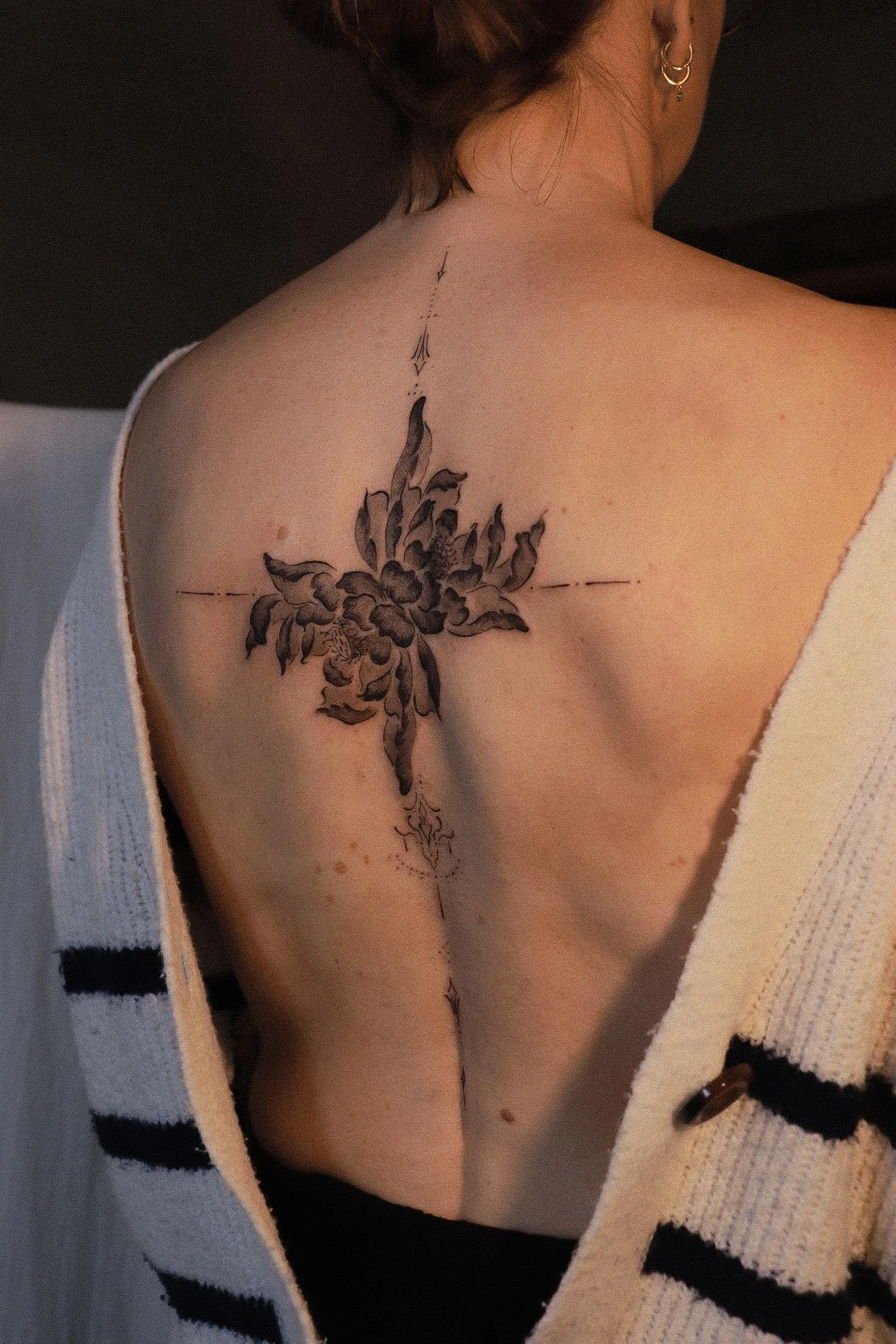 A person with short hair showing a floral tattoo on their upper chest and shoulder, with small hoop earrings in their ear, wearing a white garment with black stripes.