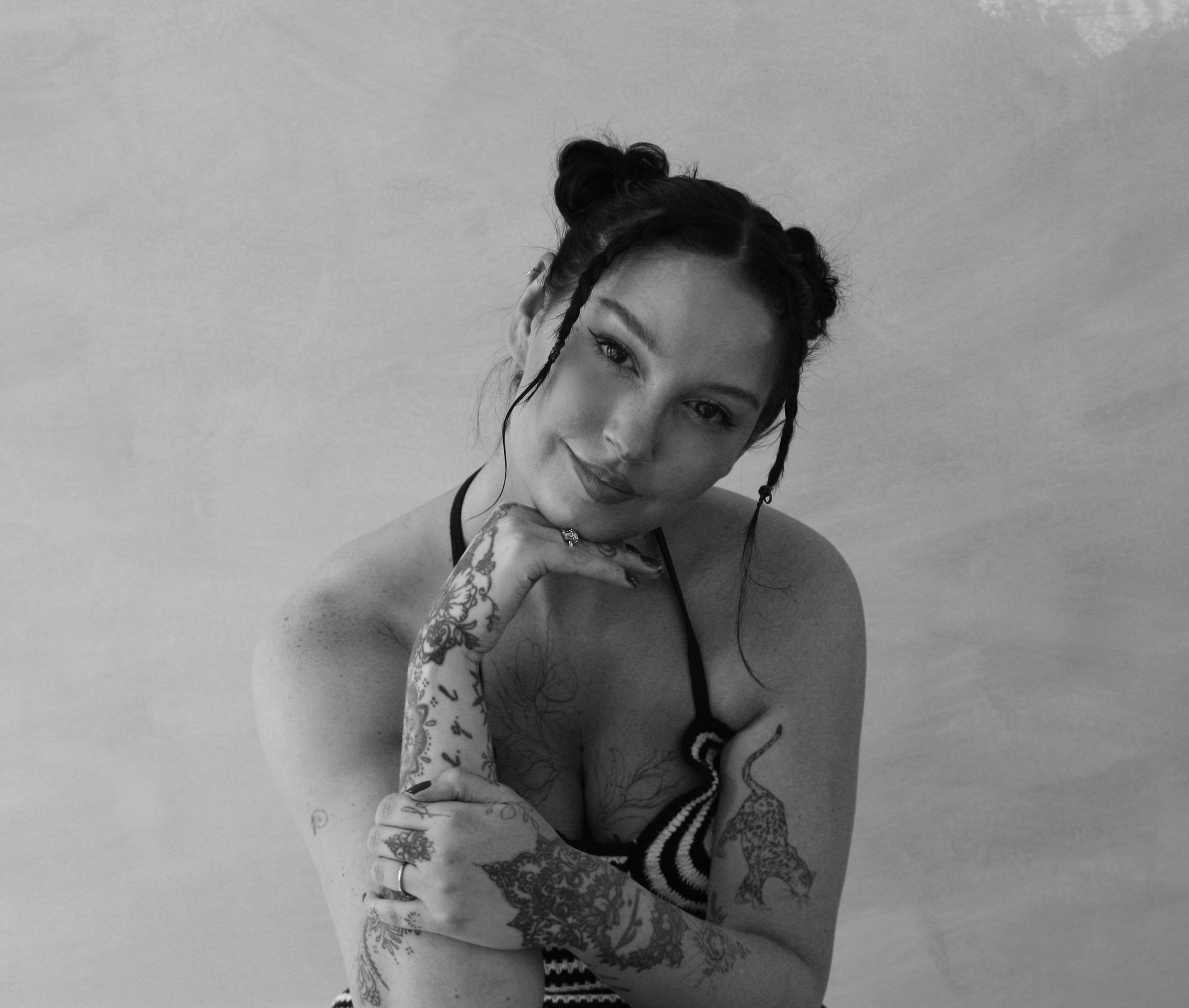 Black and white portrait of Olivia-Fayne, one of the UK's most in-demand fine line tattoo artists. With tattoos on her arms, styled hair in two buns, smiling and resting her chin on her hand.