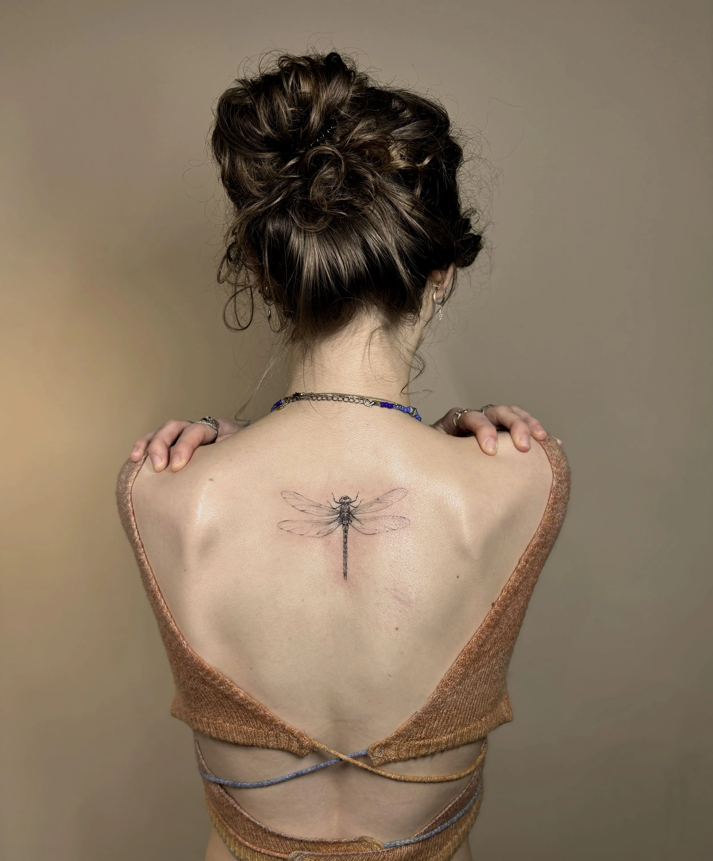 Back view of a woman with curly hair styled in a bun, wearing jewelry, and a dress with a heart-shaped top, with a tattoo of a dragonfly on her back.