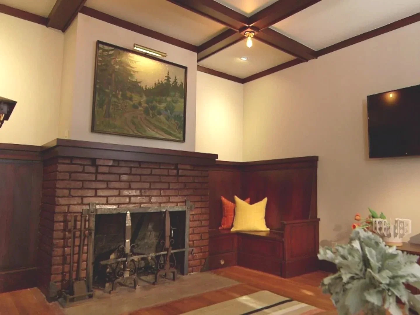 Living room with brick fireplace, wooden built-in bench with yellow and orange pillows, framed landscape painting above fireplace, flat-screen TV on the wall, white flowers in a vase, ceiling lights, wood-paneled ceiling with beams.