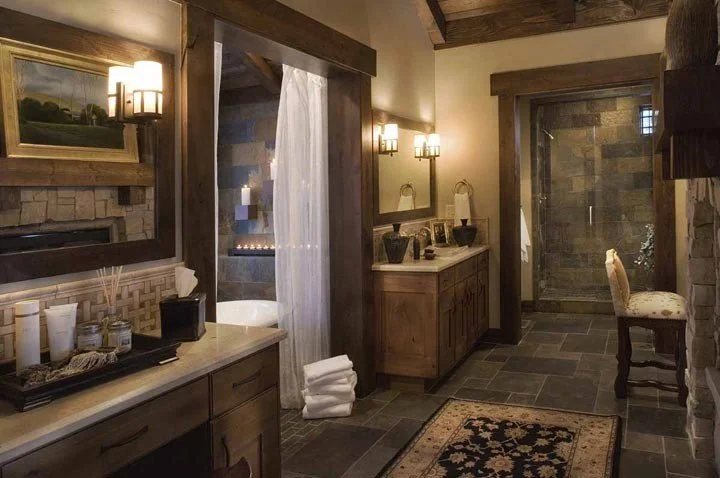 A rustic bathroom featuring stone and wooden accents, with a walk-in shower, a wooden vanity with two sinks, wall-mounted lights, a framed mirror, a small chair, and a decorative rug on a tiled floor.