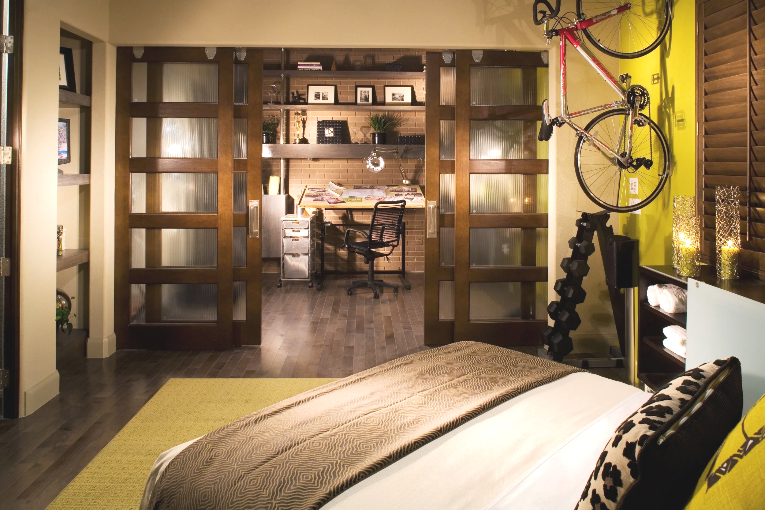 A bedroom with a bed in the foreground, and a workout station with dumbbells and a bicycle mounted on the yellow wall to the right. In the background, there is a workspace with a desk, a chair, shelves, and decorative items all behind sliding wooden and glass doors.