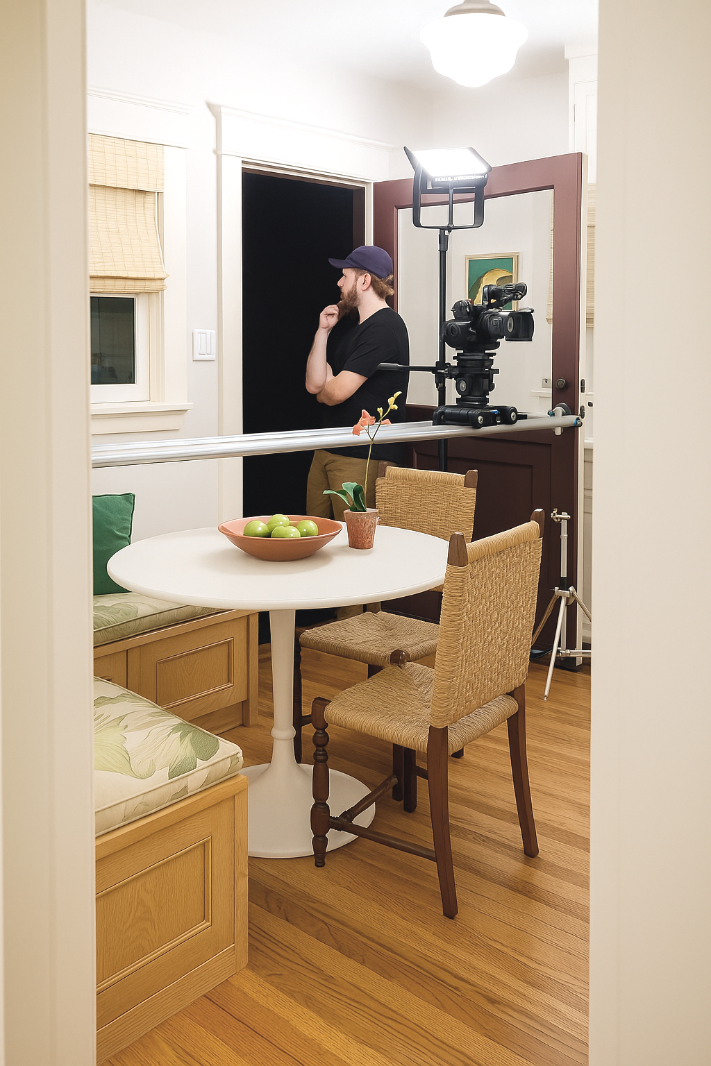 A man with a beard wearing a black T-shirt and a navy baseball cap stands with his back to a camera mounted on a sliding track, in a well-lit dining room. Behind him is a black backdrop, and there is a professional light attached to the camera setup, indicating a photo or video shoot. The dining area has a round white table with a bowl of green apples and a potted plant, wooden chairs, and a wooden bench, with hardwood floors and a window with beige blinds.